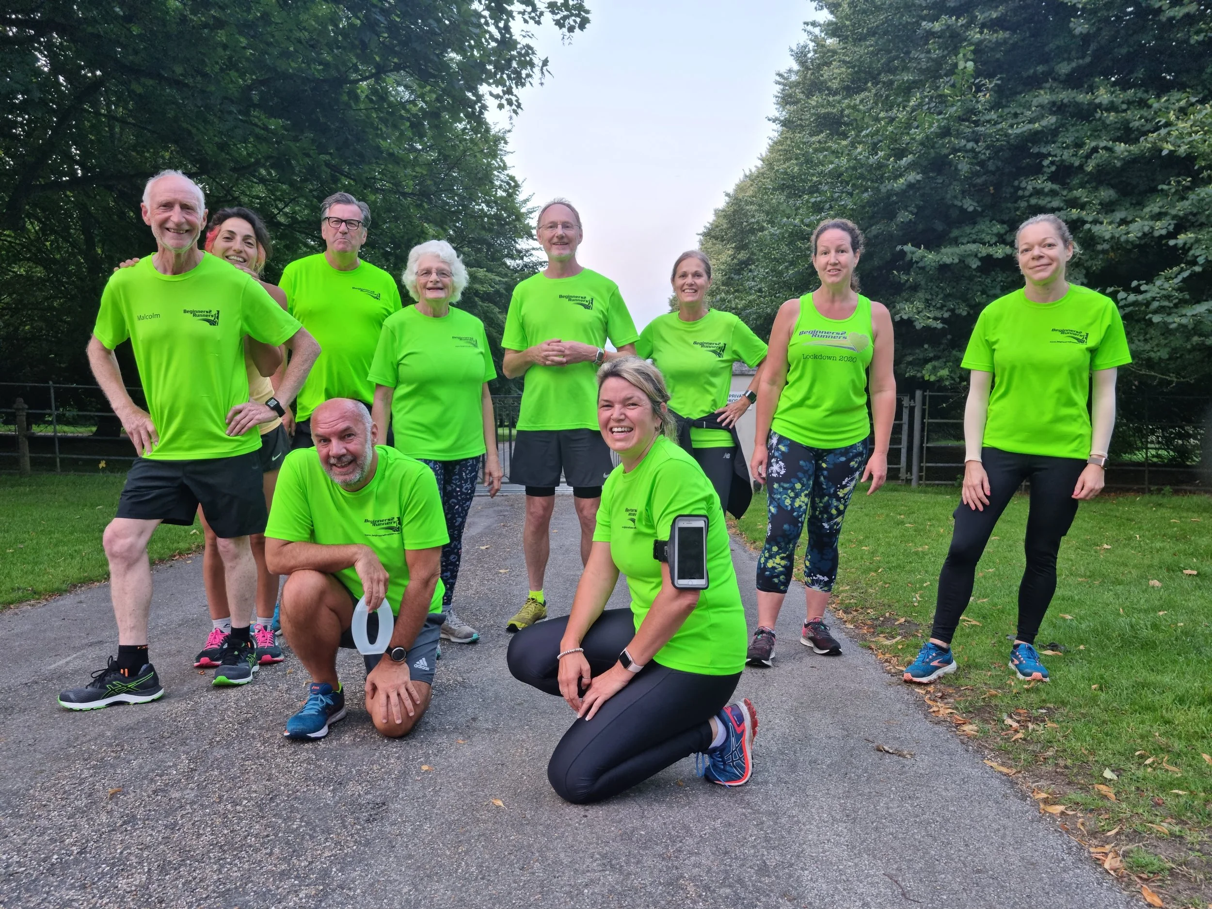 Group of people wearing matching bright green athletic shirts, gathered on a running trail in a park with green trees, smiling and posing for a photo after a workout.