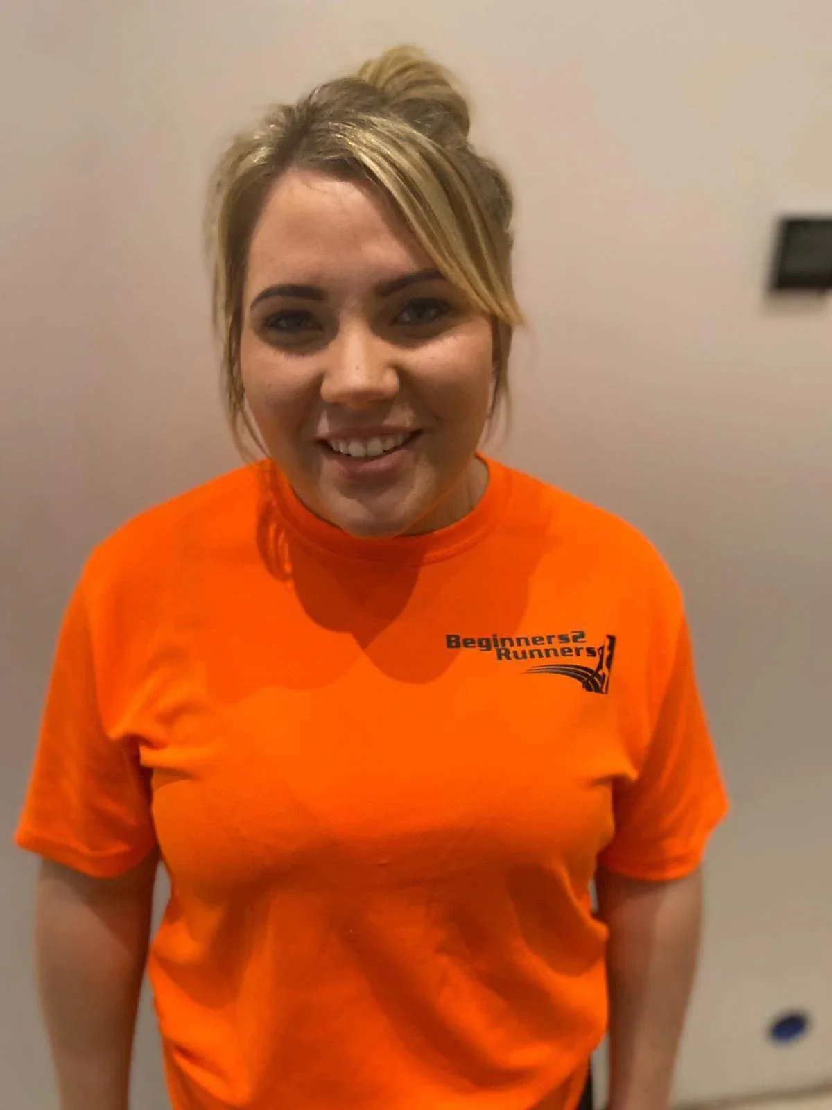 A young woman with blonde hair in a messy bun, smiling and wearing an orange club top that says 'Beginners 2 Runners'.