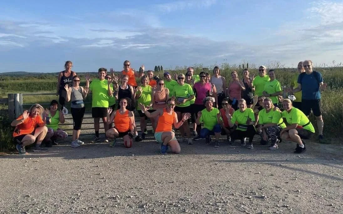 Group of B2R members participating in an outdoor fitness event or running club, many wearing bright club tops, posing on a gravel path with a rural landscape in the background.