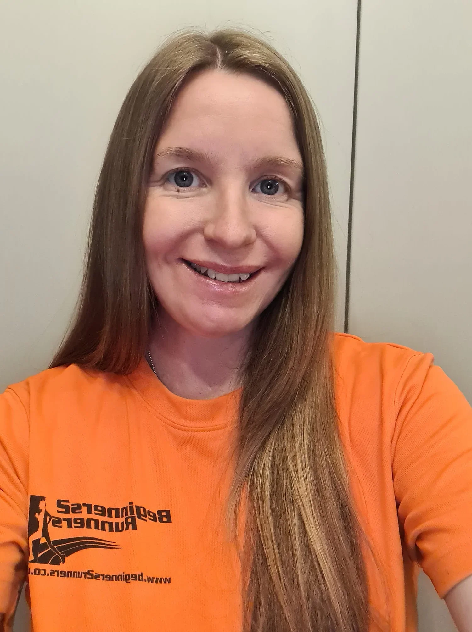 Woman with long reddish-brown hair smiling, wearing an orange t-shirt with black text and logo, in front of a plain wall.