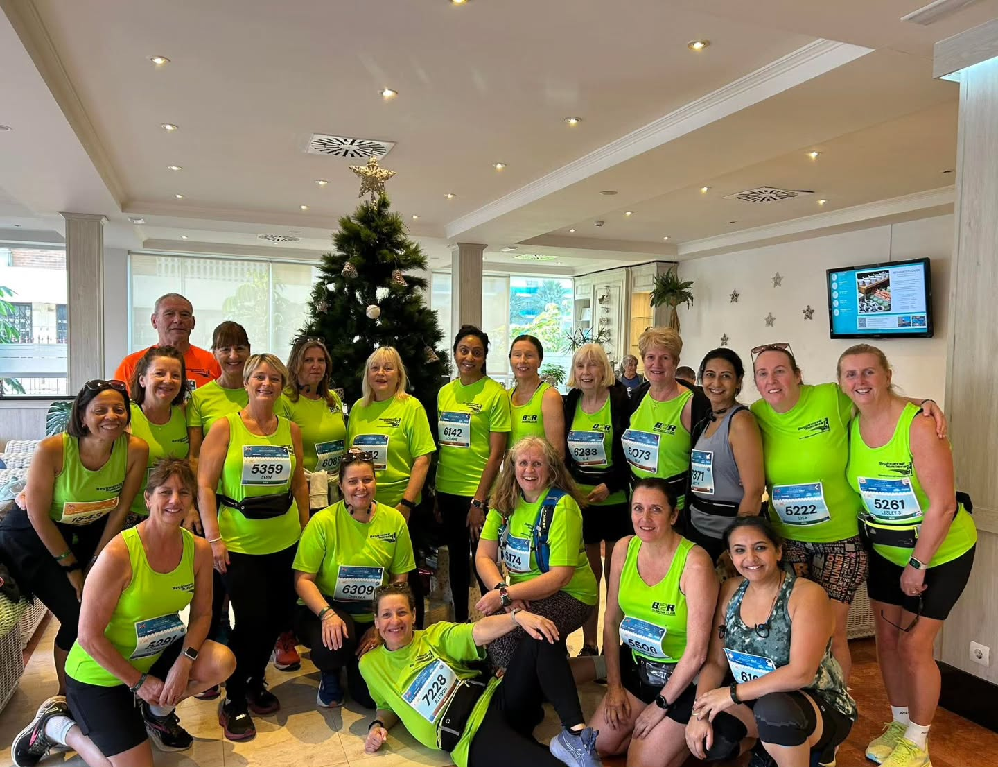 Group of B2R runners in neon green club tops gathered indoors in front of a decorated Christmas tree before a running event in Benidorm.