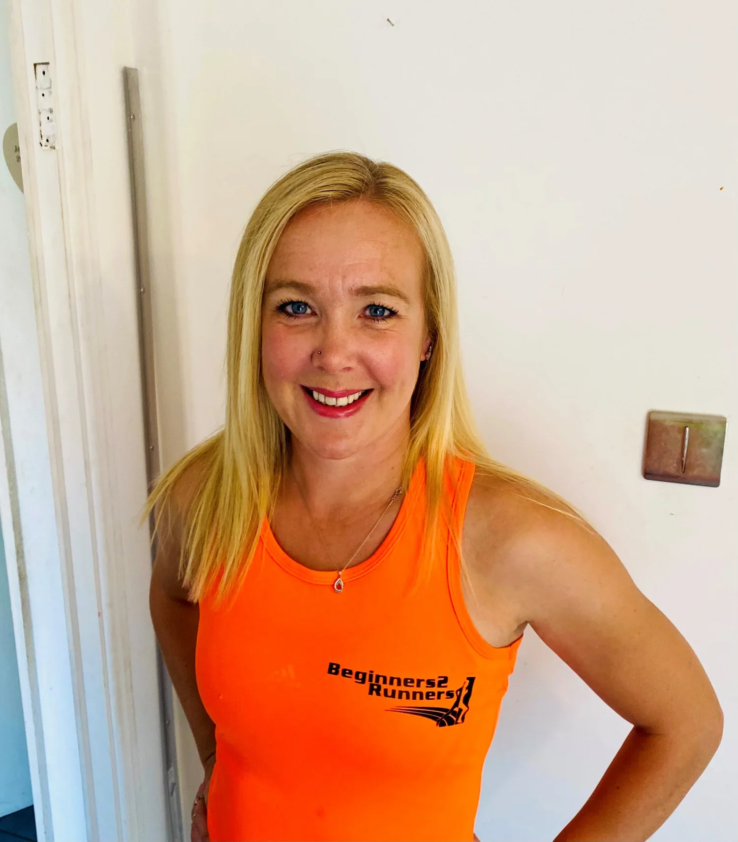 A smiling blonde woman with blue eyes wearing an orange club vest with 'Beginners2 Runners!' printed on it, standing indoors near a white wall.