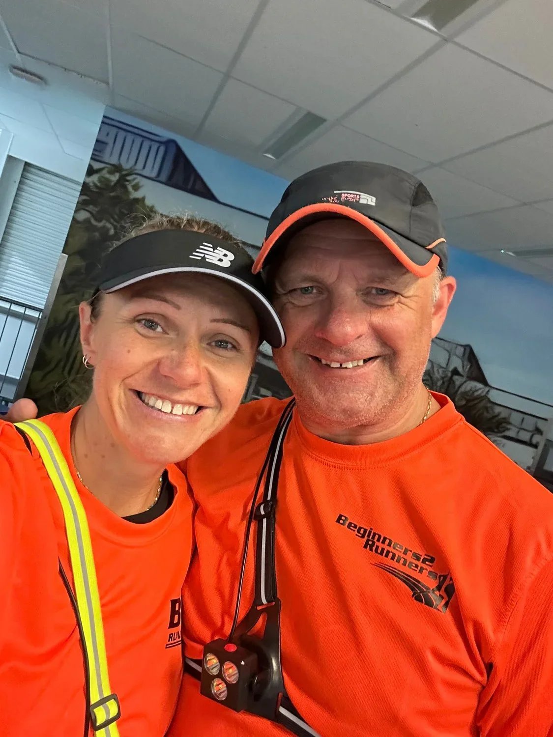 A smiling man and woman taking a selfie together indoors, both wearing orange club tops with B2R related logos and caps, with the woman also wearing a black and silver New Balance visor.
