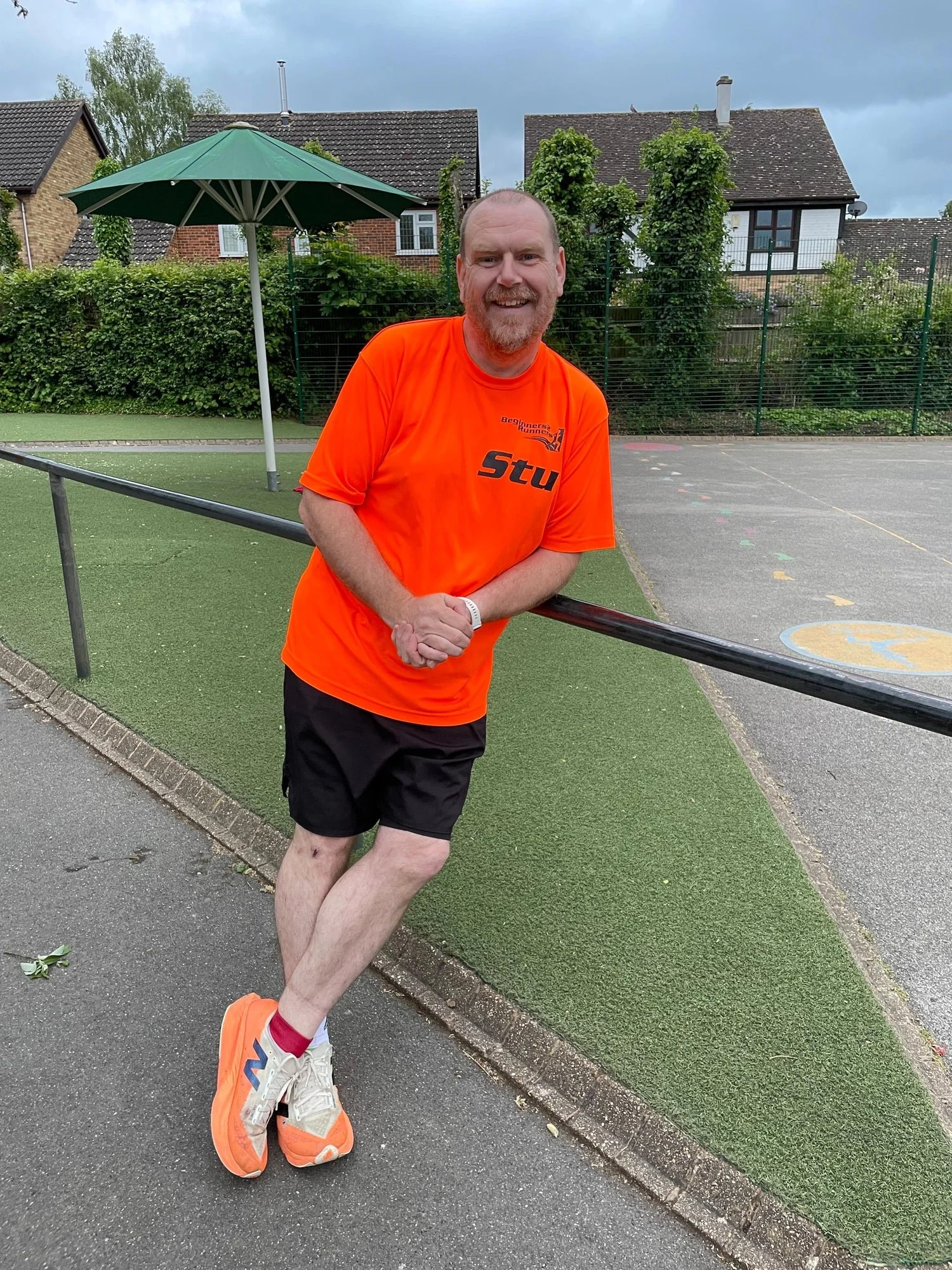 A man with short hair and a beard, wearing an orange shirt, black shorts, and orange running shoes, standing on a sidewalk near a sports court, leaning against a black railing, smiling at the camera.