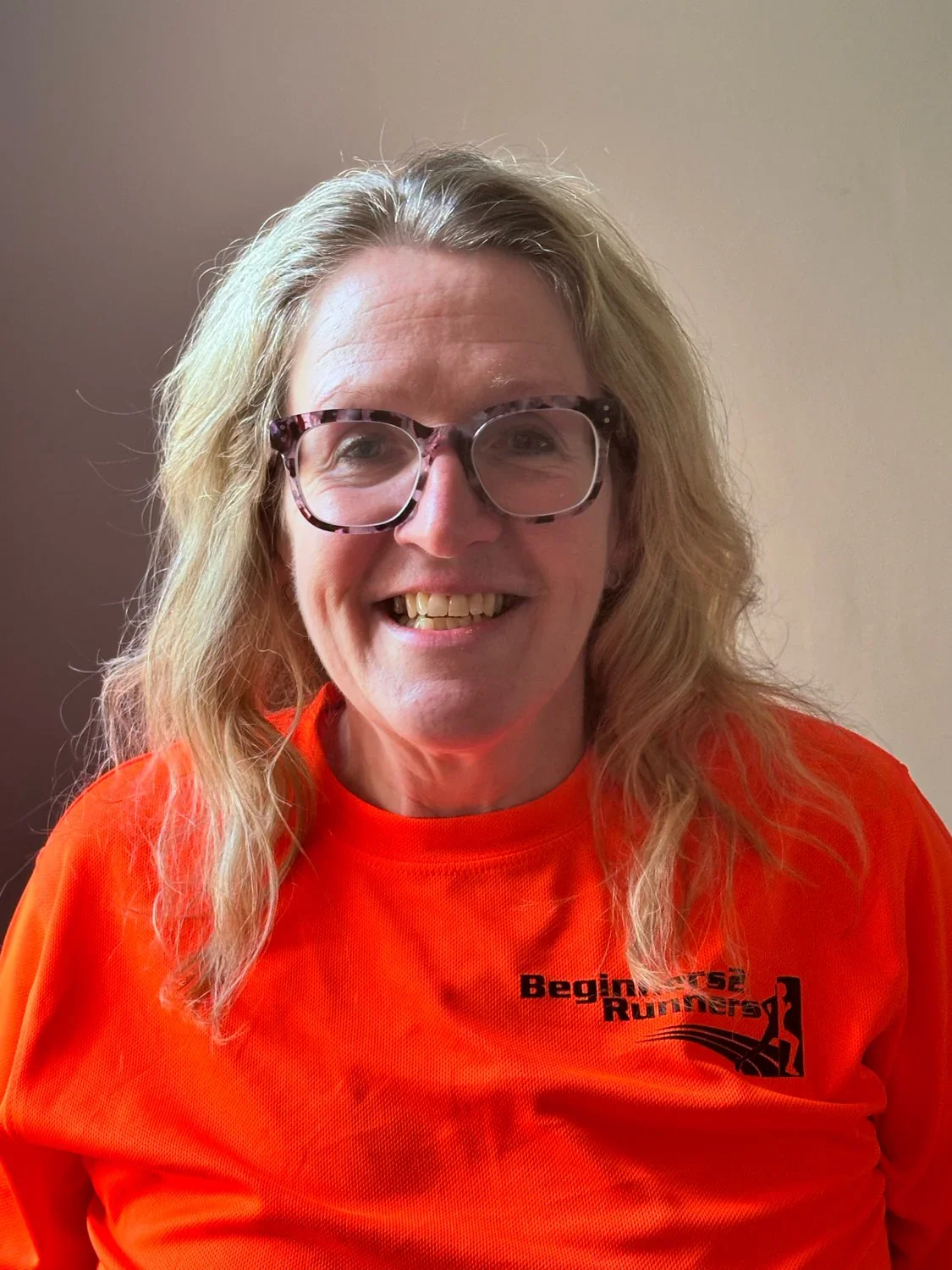A woman with blonde hair, wearing large glasses and an orange athletic shirt with the text 'Beginner 52 Runner' and a running figure logo, smiling at the camera.