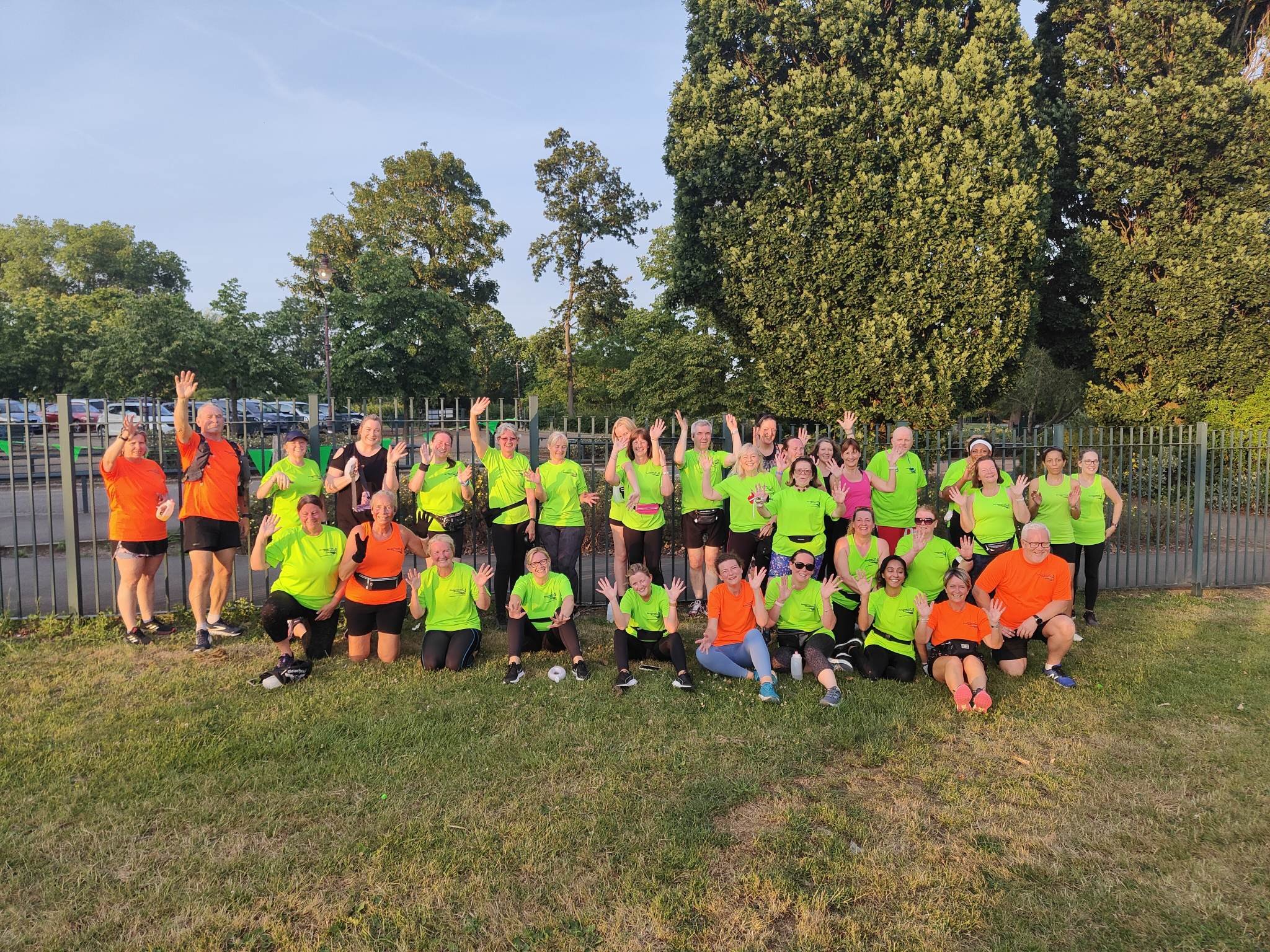 Group of people in bright green and orange athletic shirts, posing outdoors on grass, waving and smiling, with trees and a fence in the background.