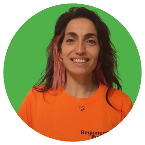 A woman with wavy black and pink hair smiling, wearing an orange club top and a necklace with a cloud pendant, against a green circular background.