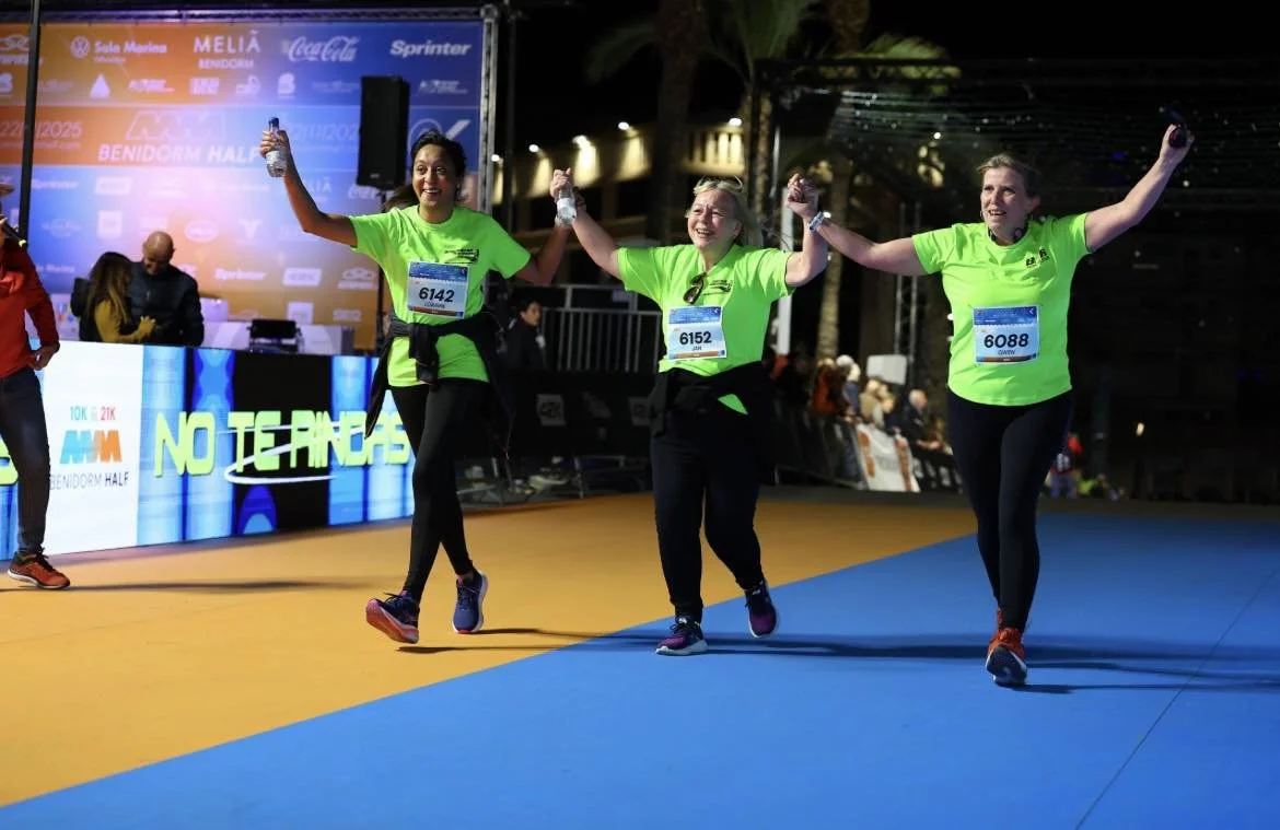 Three women in bright green shirts holding hands and celebrating on a running track at a marathon event, with a digital display board in the background.