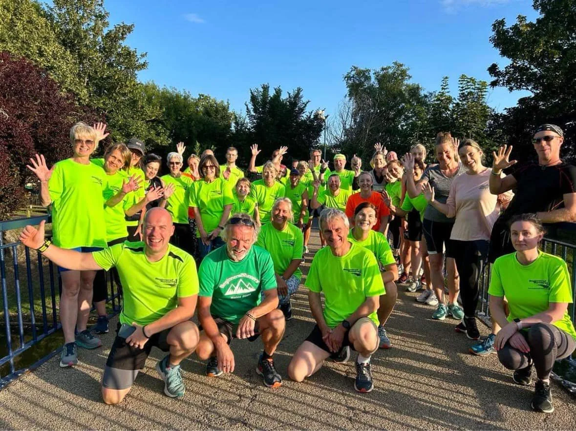 Group of people in bright yellow and green athletic shirts, smiling and waving, outdoors on a sunny day near a pond with trees in the background.