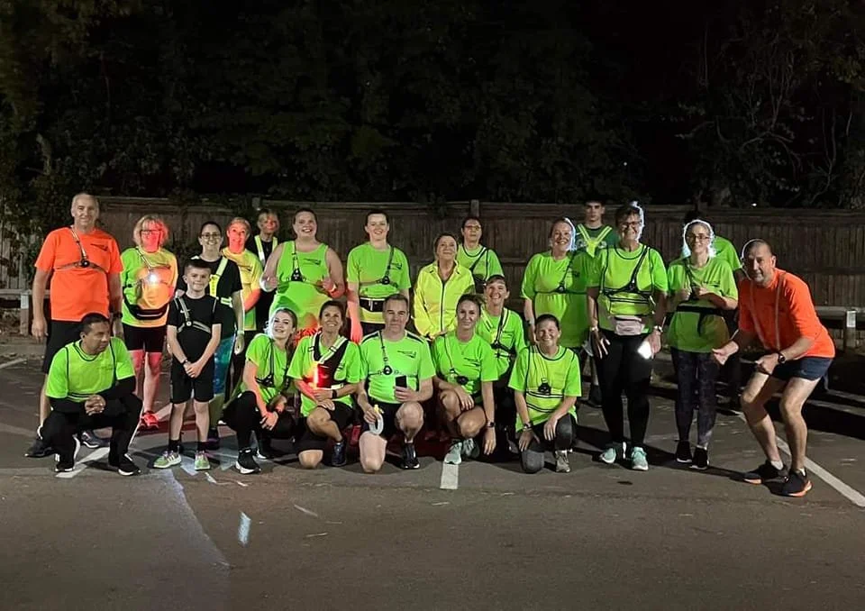 A group of 19 B2R members, most wearing club tops and running lights, posing for a photo at night outdoors. The background includes a wooden fence and trees.