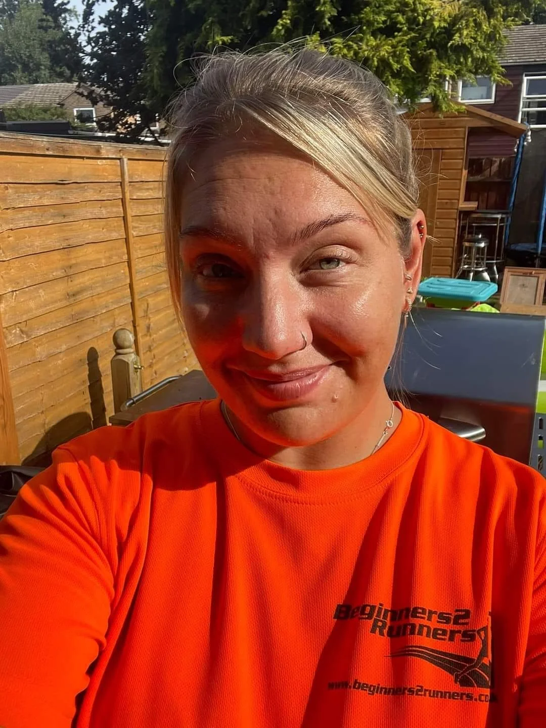 A woman taking a selfie outdoors in sunlight, wearing an orange shirt that says 'Beginners Runners'. She has blonde hair, blue eyes, and piercings on her nose and ear, with a cheerful expression.