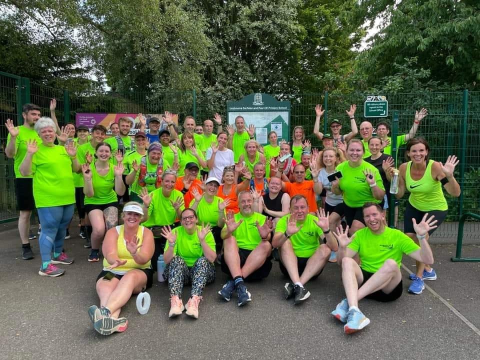 Group of people in bright athletic clothing, mostly neon green, posing outdoors after a run or walk, with arms raised and smiling.