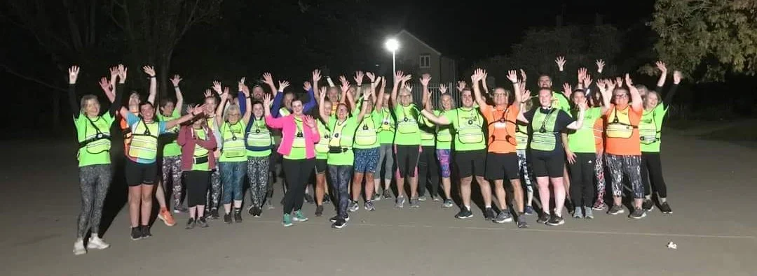 Group of people wearing bright neon club tops, raising their hands in the air during a nighttime club session.