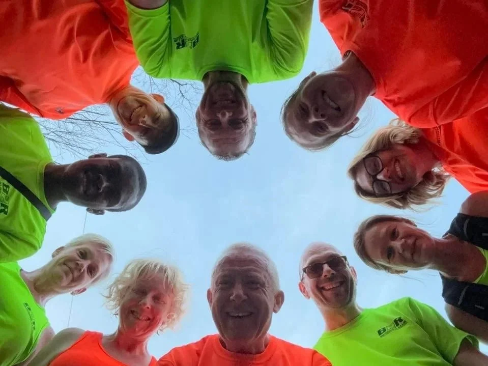 Group of smiling people standing in a circle and looking down at the camera, outside on a clear day.