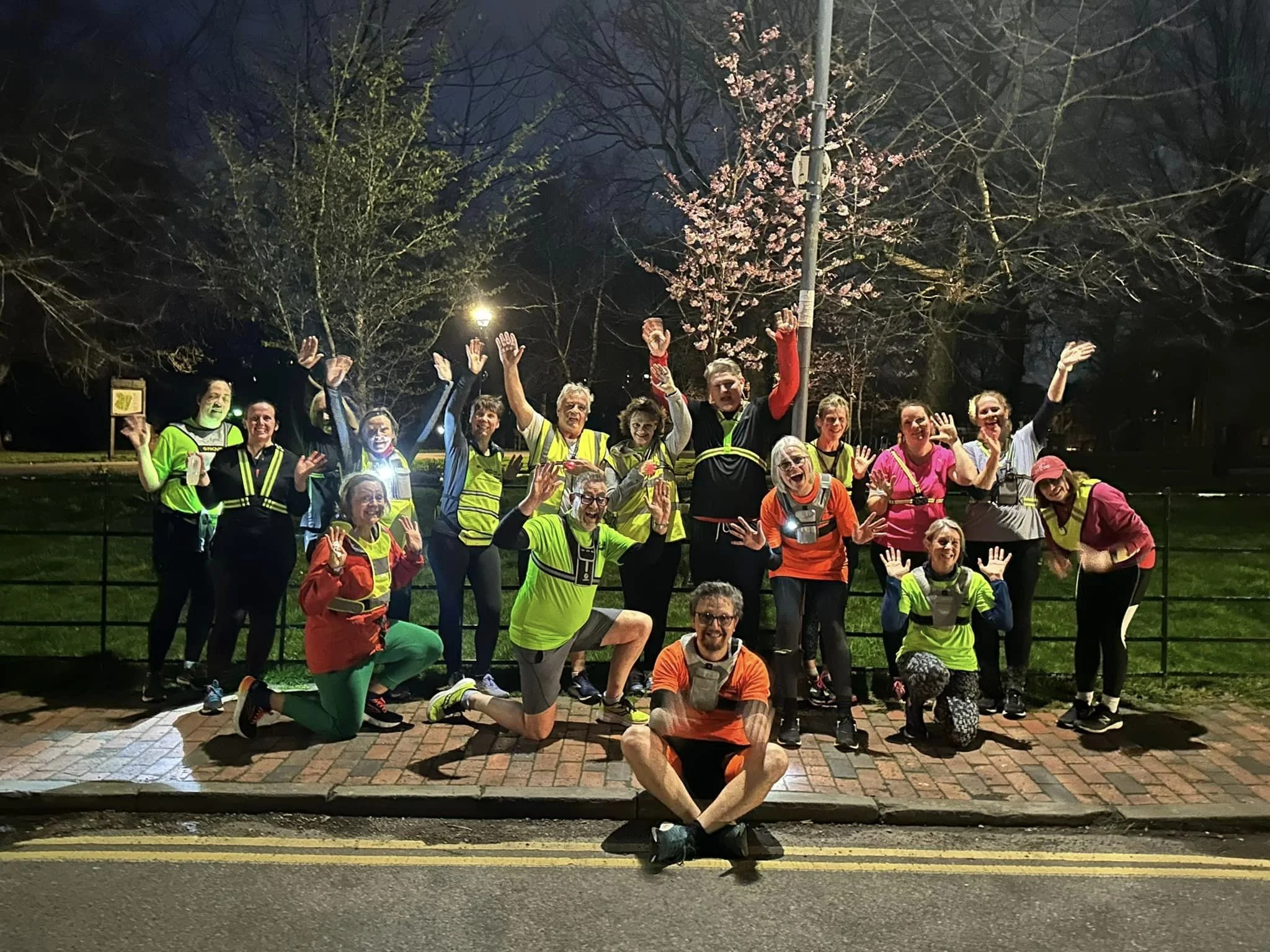 Group of B2R members dressed in running or athletic gear, posing with their hands raised, outdoors at night, some wearing reflective vests, with leafless trees and a flowering tree in the background.