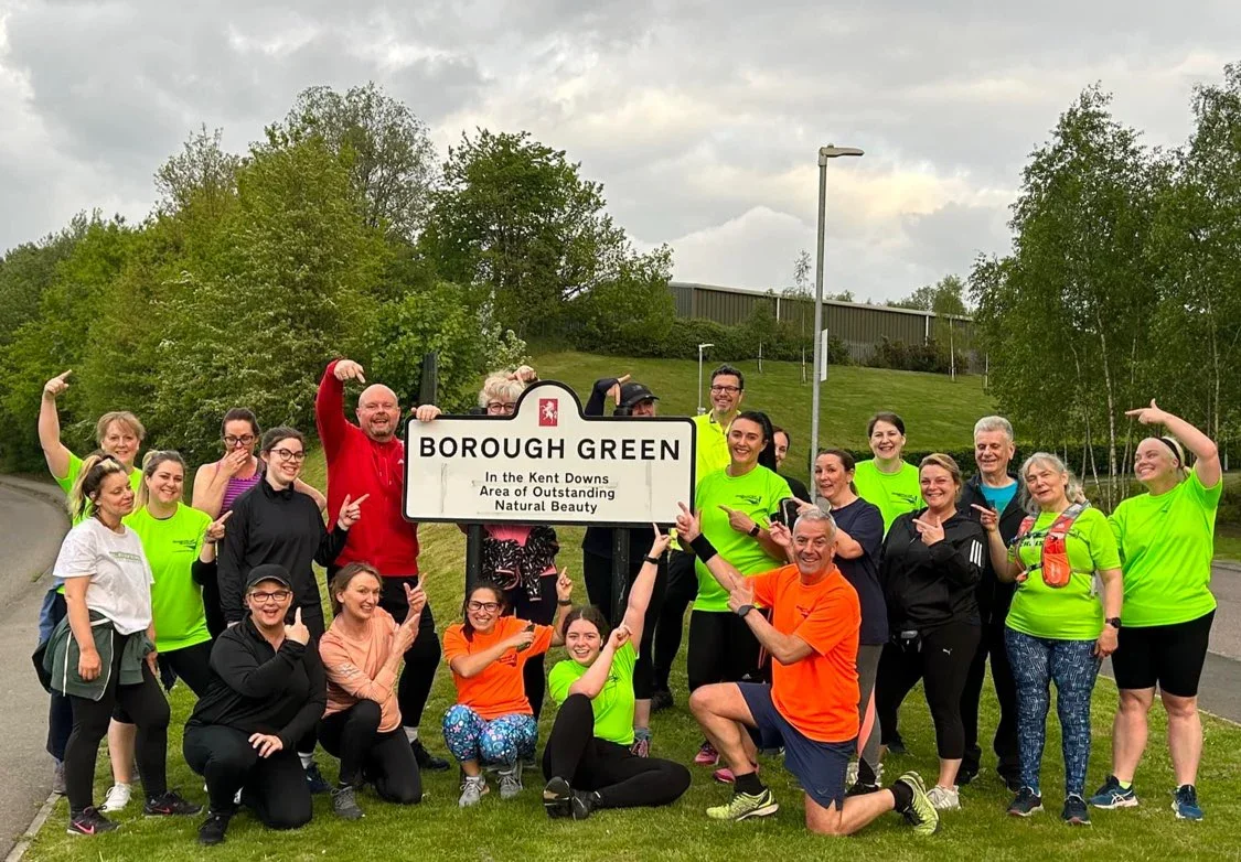 A group of people gathered outdoors in front of a sign that reads "Borough Green" and "In the Kent Downs Area of Outstanding Natural Beauty." They are smiling and pointing at the sign, some making flexing gestures. The group includes men and women of various ages, some wearing bright green shirts, and the background features trees, grass, a road, and cloudy sky.