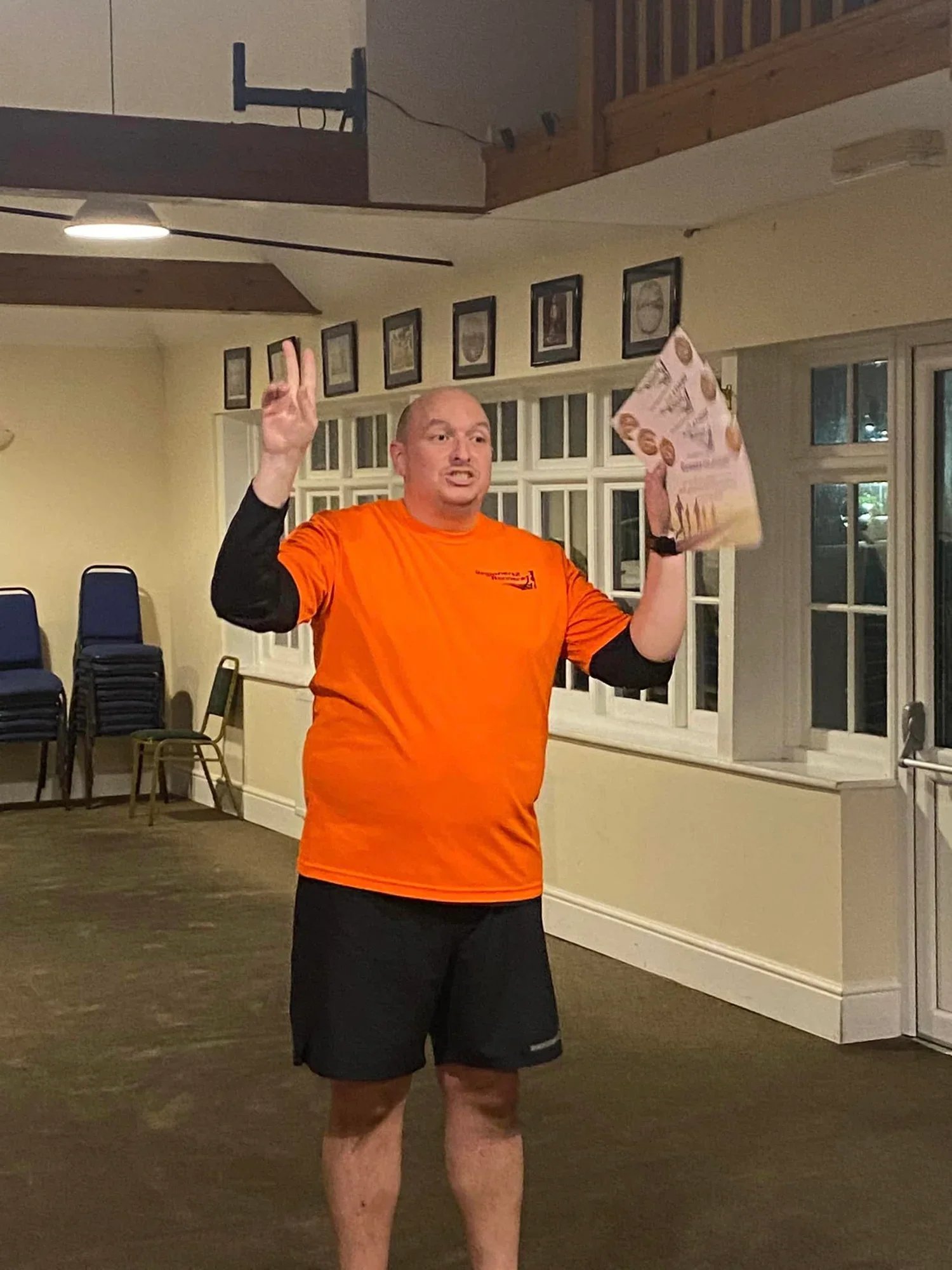 Man in an orange club top and black shorts, in a room with stacked chairs and pictures on the wall.