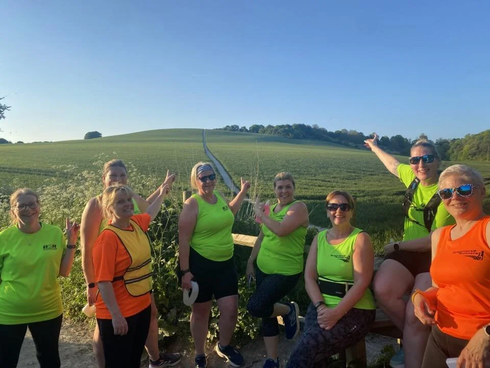 Group of and women in neon green & orange cub tops posing outdoors on a trail with a green grassy hill and blue sky in the background.