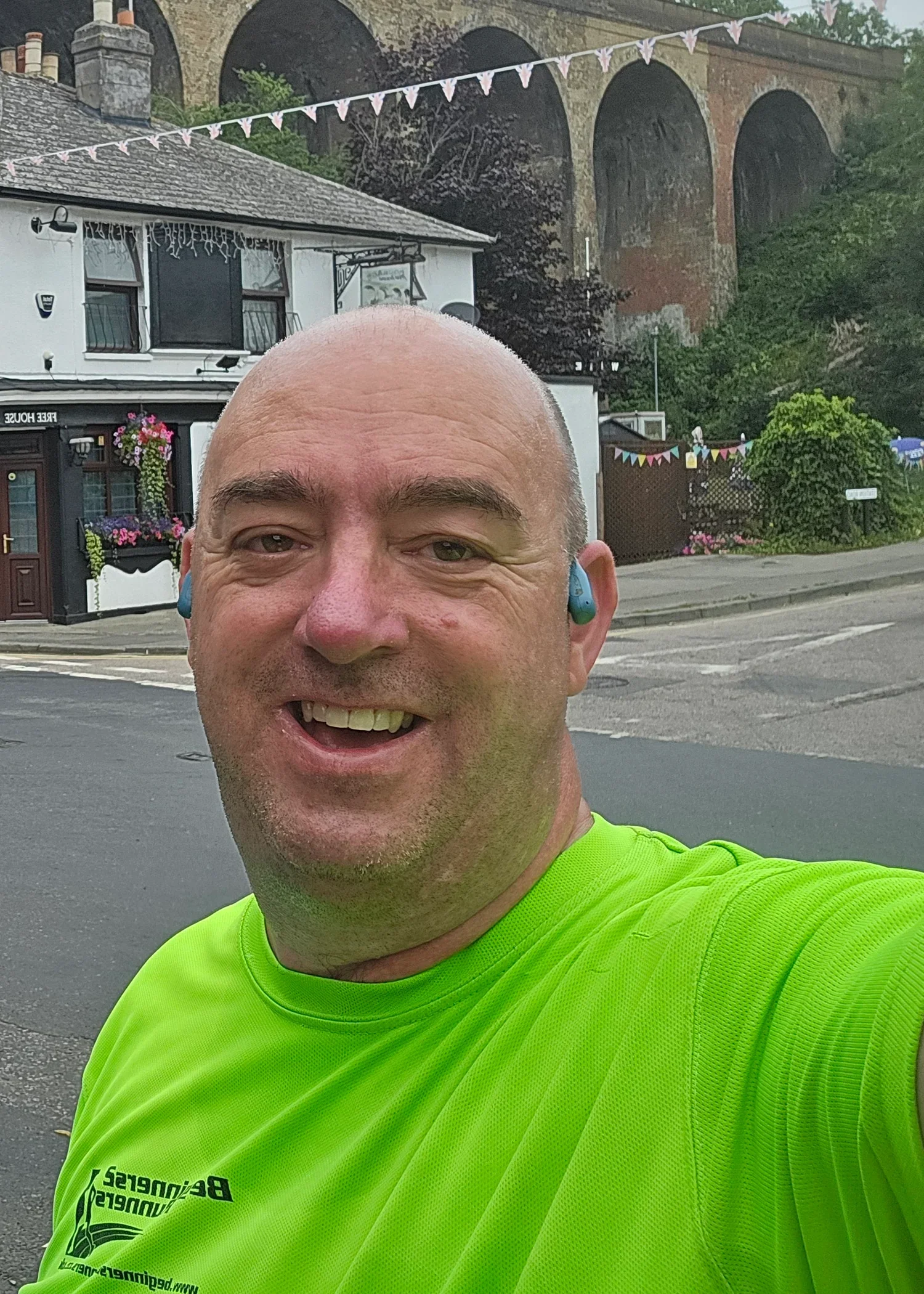 A man smiling and wearing a bright green clb top and blue earbuds, taking a selfie outdoors on a street.