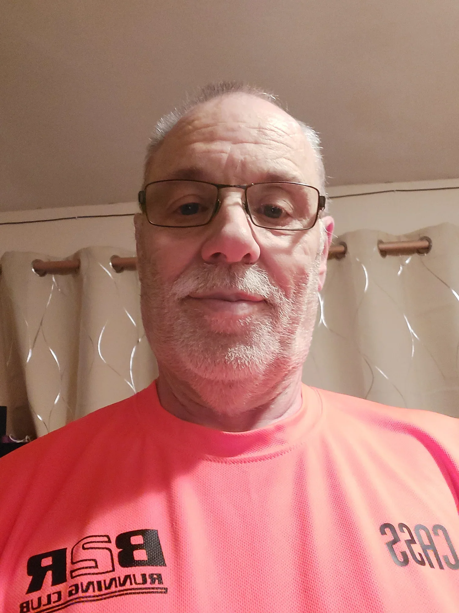 A middle-aged man with glasses, white beard, and short hair wearing an orange club top that says 'B2R Running Club' on it. He is indoors.