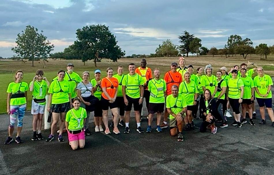 Group of people wearing bright club tops, posing outdoors on a paved area with trees and grassy fields in the background.