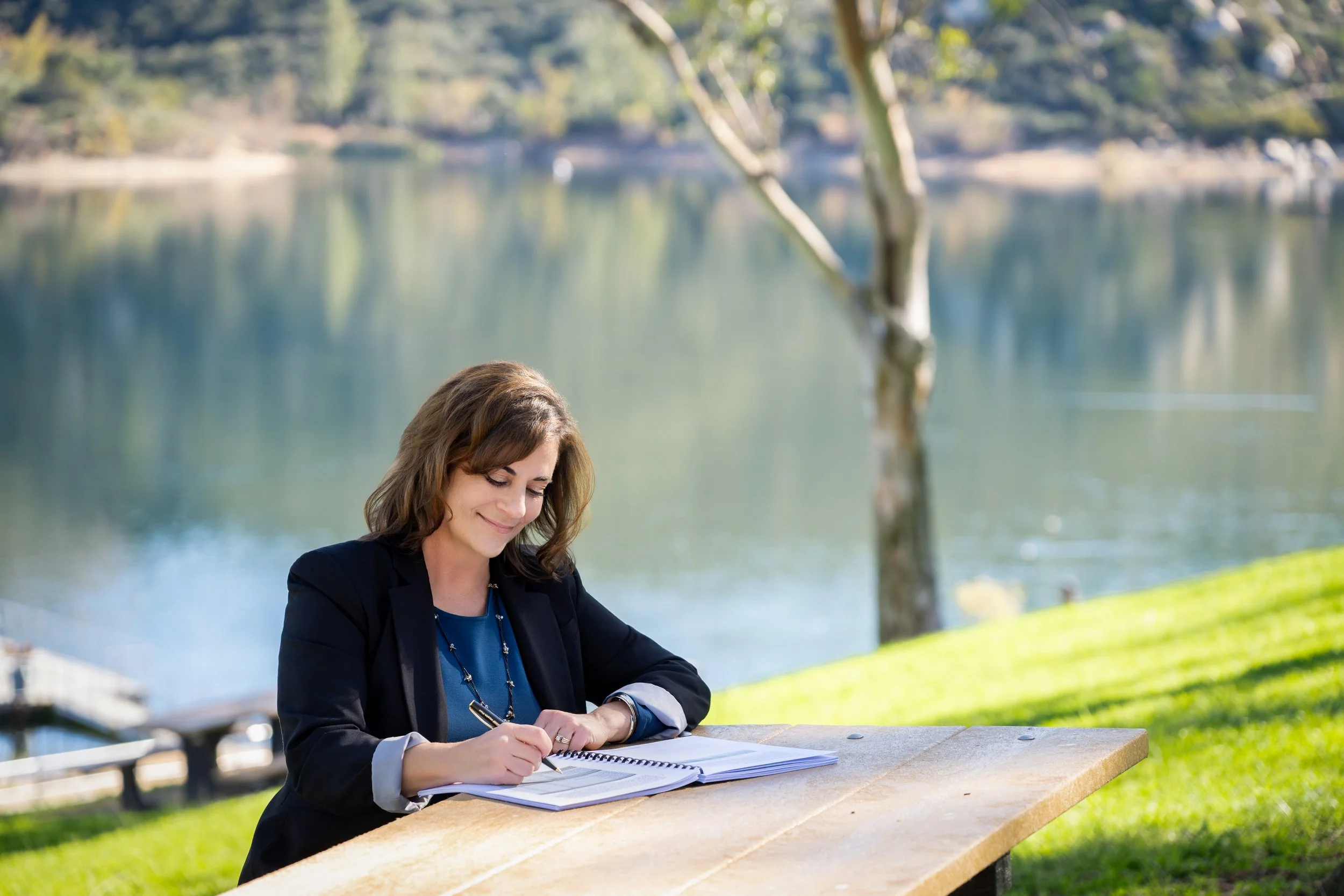 A woman sitting at a wooden picnic table outdoors, writing in a notebook, with a lake and trees in the background.