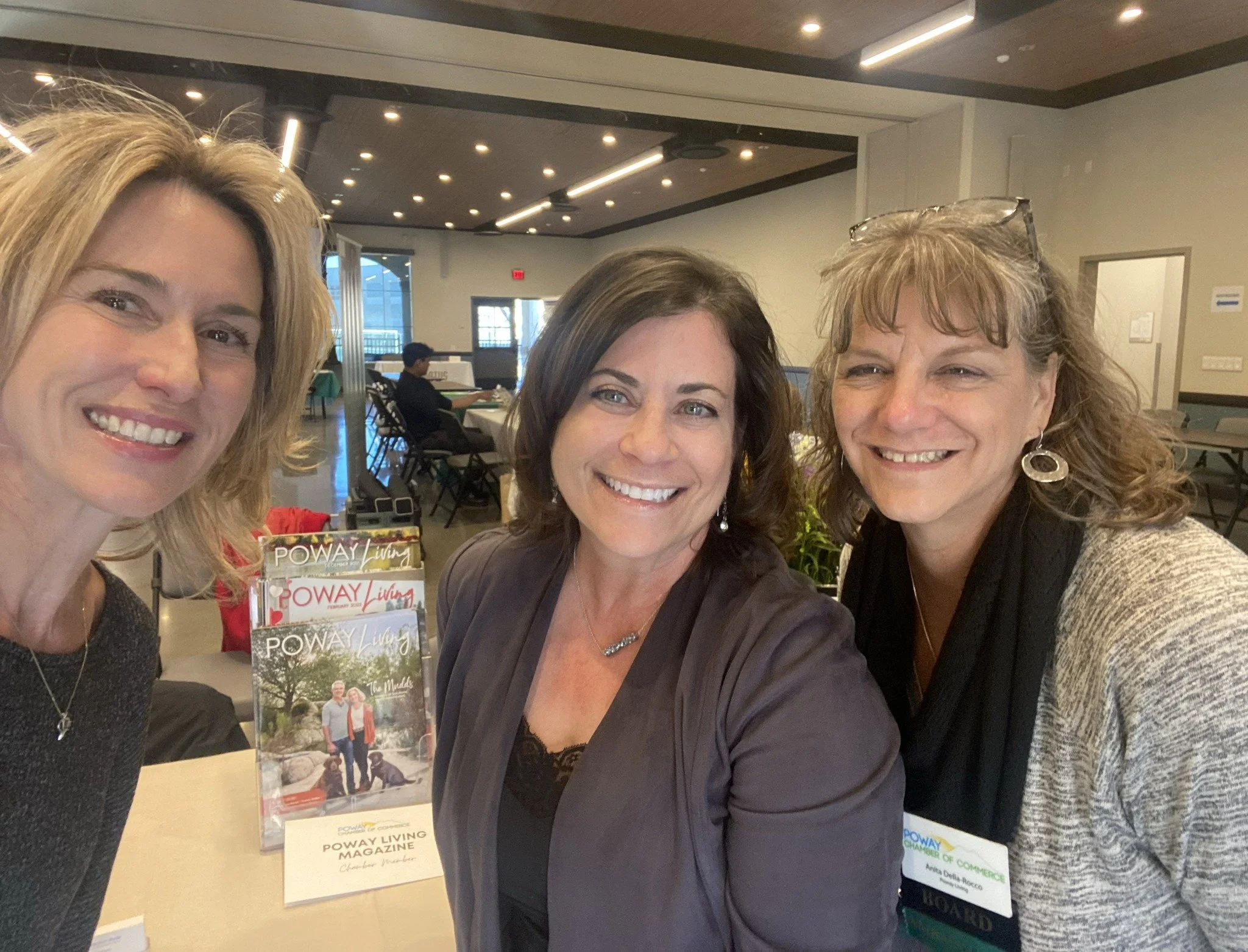 Three women smiling at a table during an event, with magazines and name tags visible. One woman wears a black top, the second a dark blazer, and the third a light-colored cardigan. The background shows other attendees in a large, well-lit room.