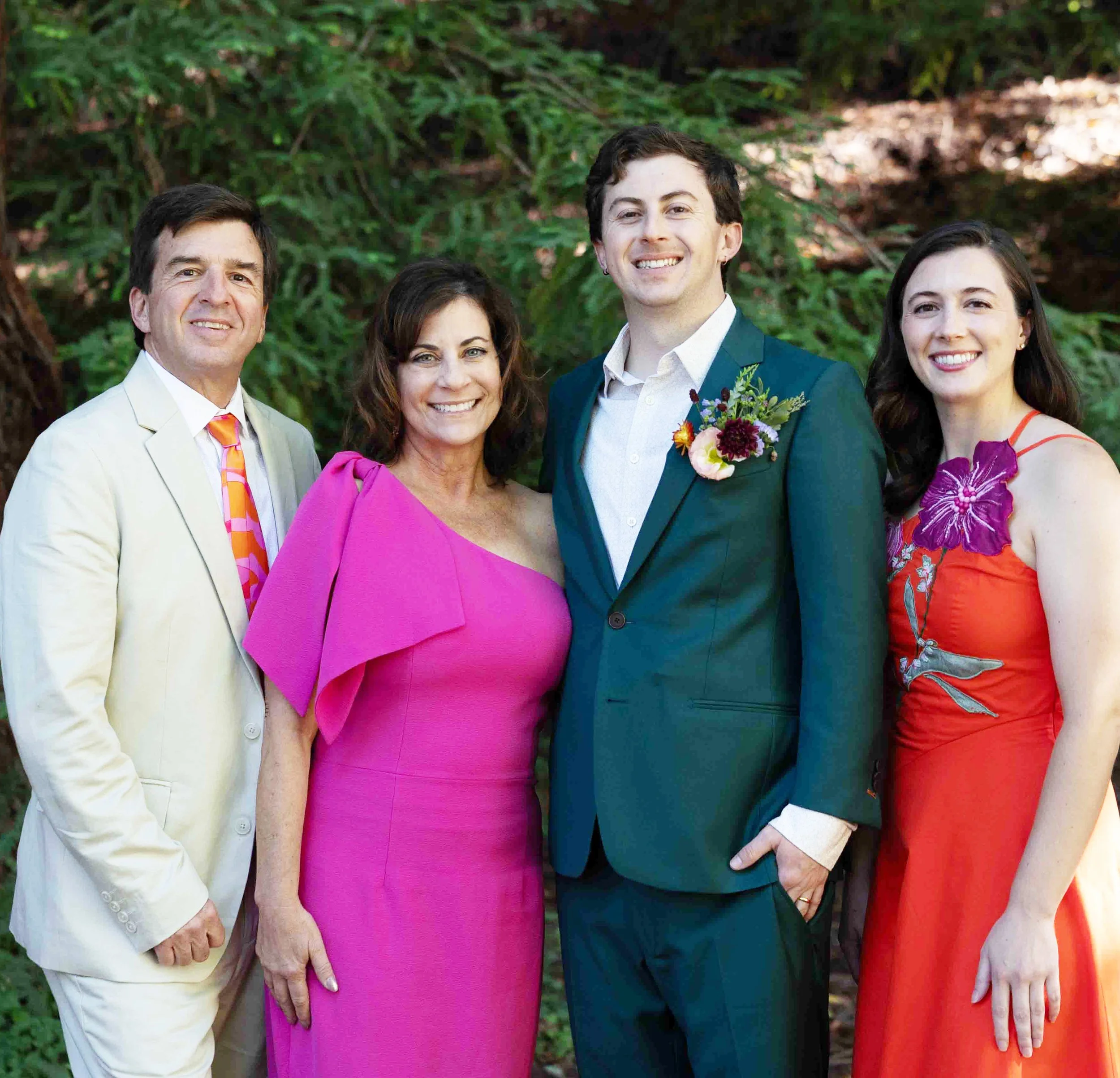A group of four people, two men and two women, dressed in formal attire, standing outdoors with a green, leafy background, smiling for the camera.