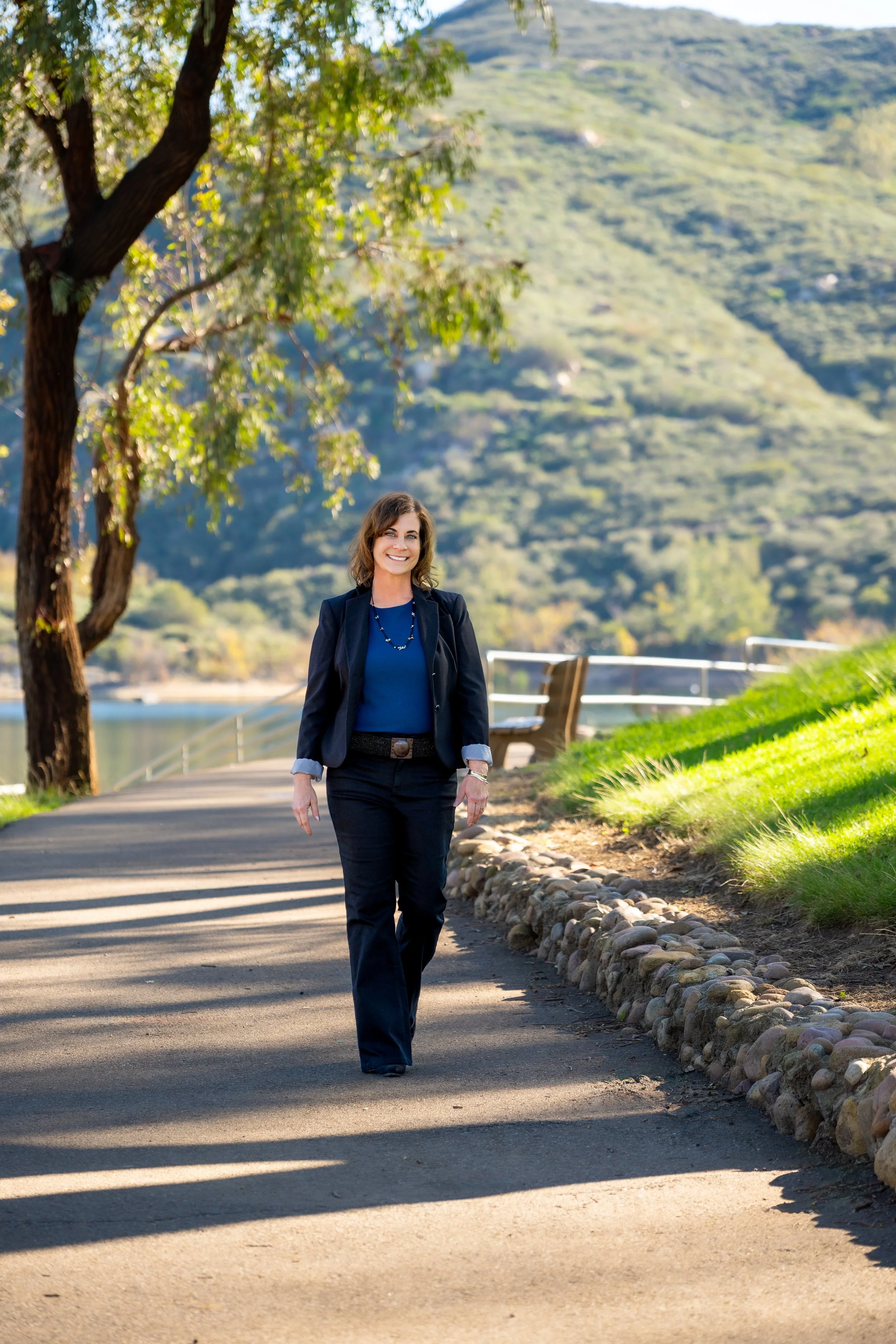 A woman in business attire walking along a park pathway with trees, grass, and a mountain in the background.