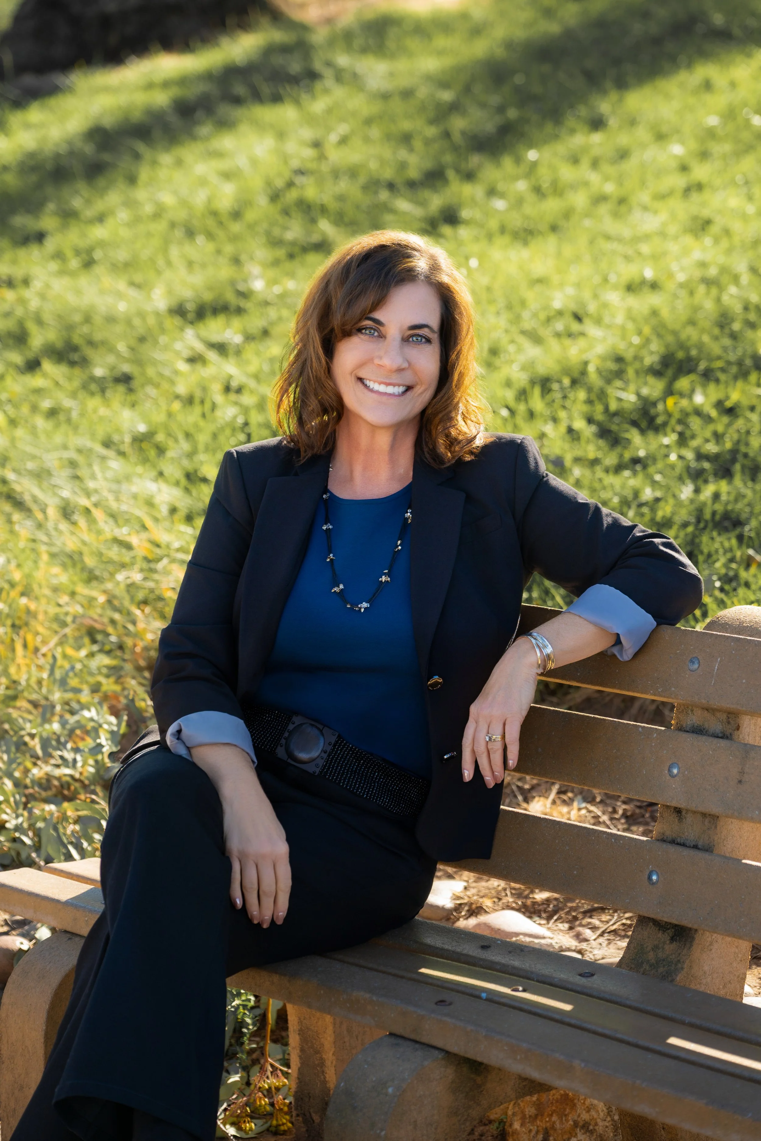 A woman sitting on a park bench smiling at the camera, wearing a dark blazer and blue top, with a green grassy background.