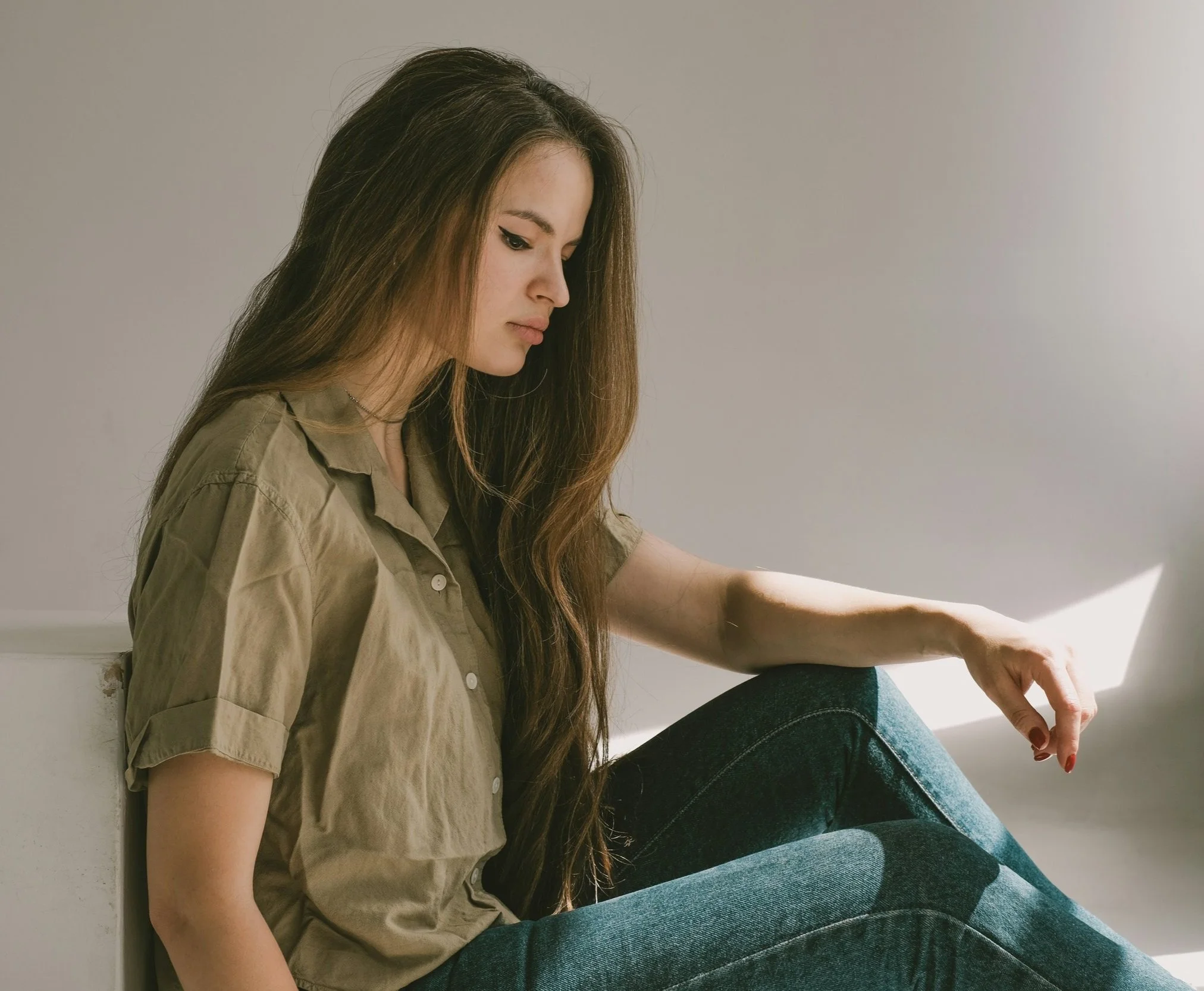 A young woman with long brown hair sitting on a white bench, looking down thoughtfully, wearing a khaki button-up shirt and blue jeans in a minimal plain room.
