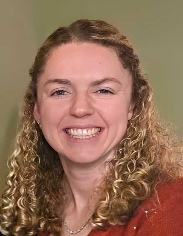 A woman with curly blonde hair smiling in front of a plain background.