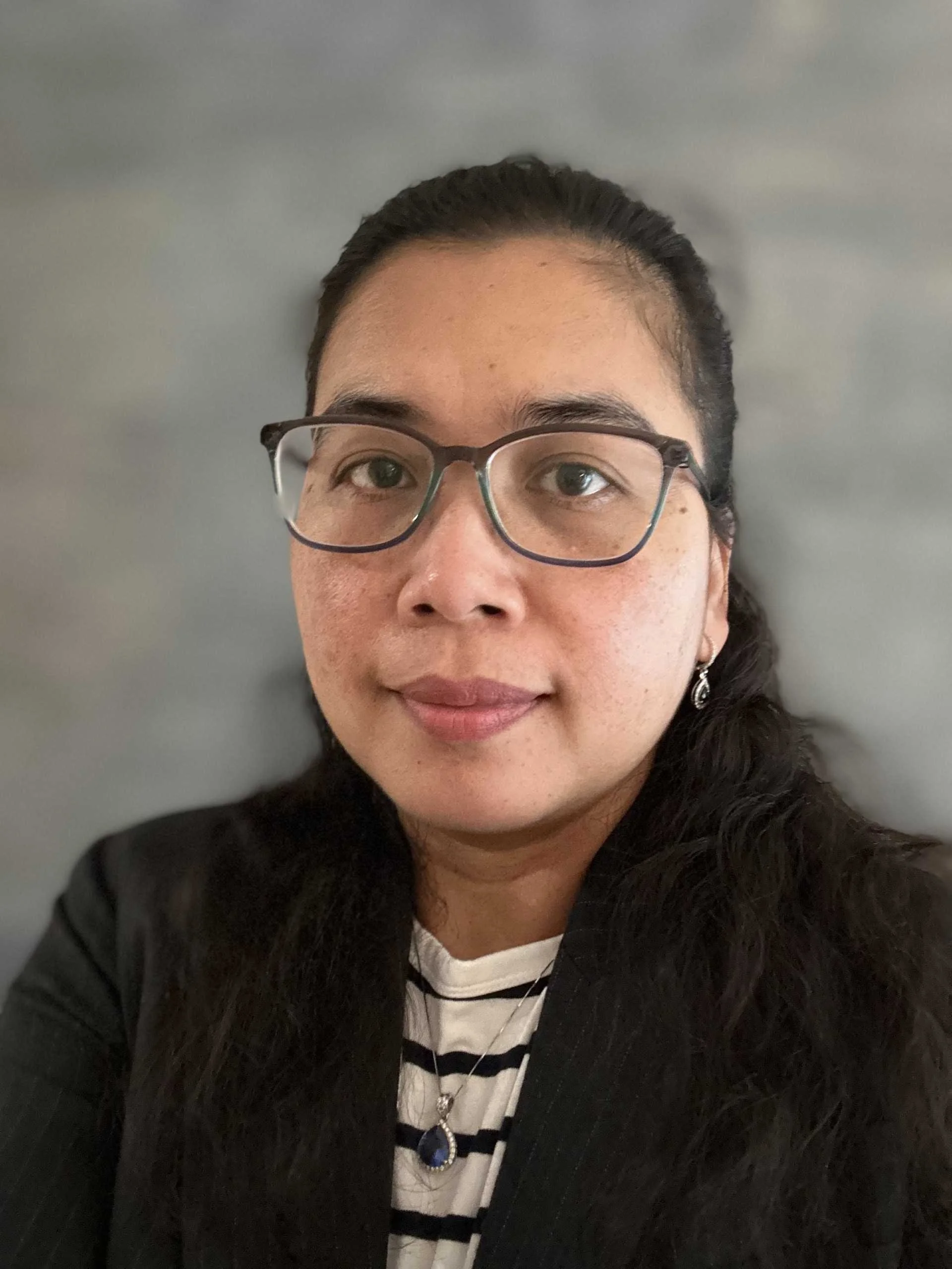 A woman with long dark hair, glasses, and earrings, wearing a black blazer over a striped shirt, smiling slightly, against a plain gray background.
