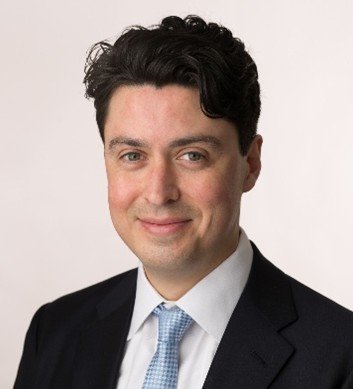 Portrait of a man in a suit and tie, with dark curly hair, smiling at the camera.
