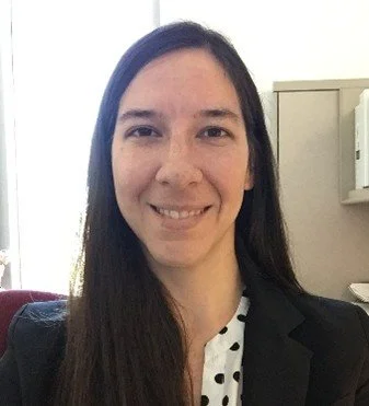 A woman with long dark hair smiling, wearing a black blazer and a white shirt with black polka dots, indoors with a light-colored wall and a cabinet behind her.