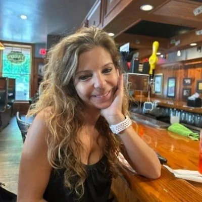 A woman with long curly hair, smiling and resting her head on her hand at a bar or restaurant. The bar counter is visible with some objects on it, and the background shows a wooden interior and large windows.