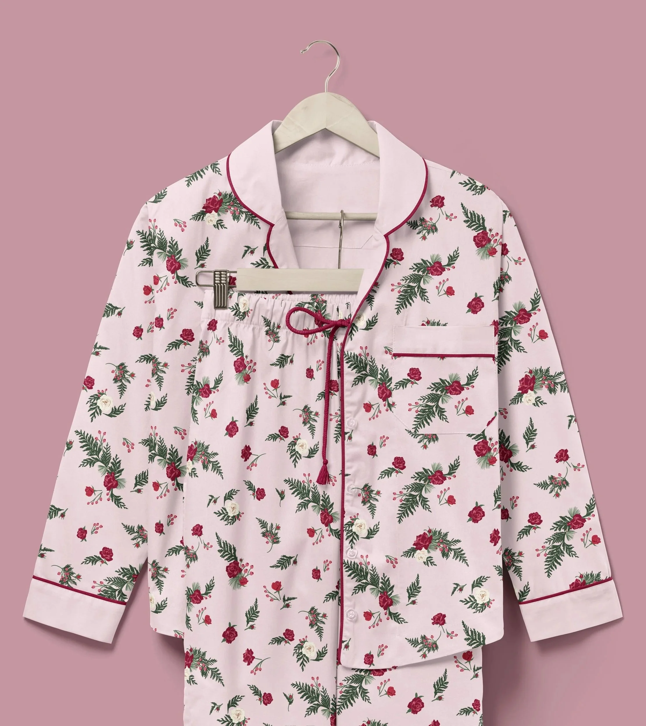 Pajama set in soft blush pink printed with scattered winter roses, evergreen sprigs, berries, and pinecones, finished with cranberry piping and ribbon trim.