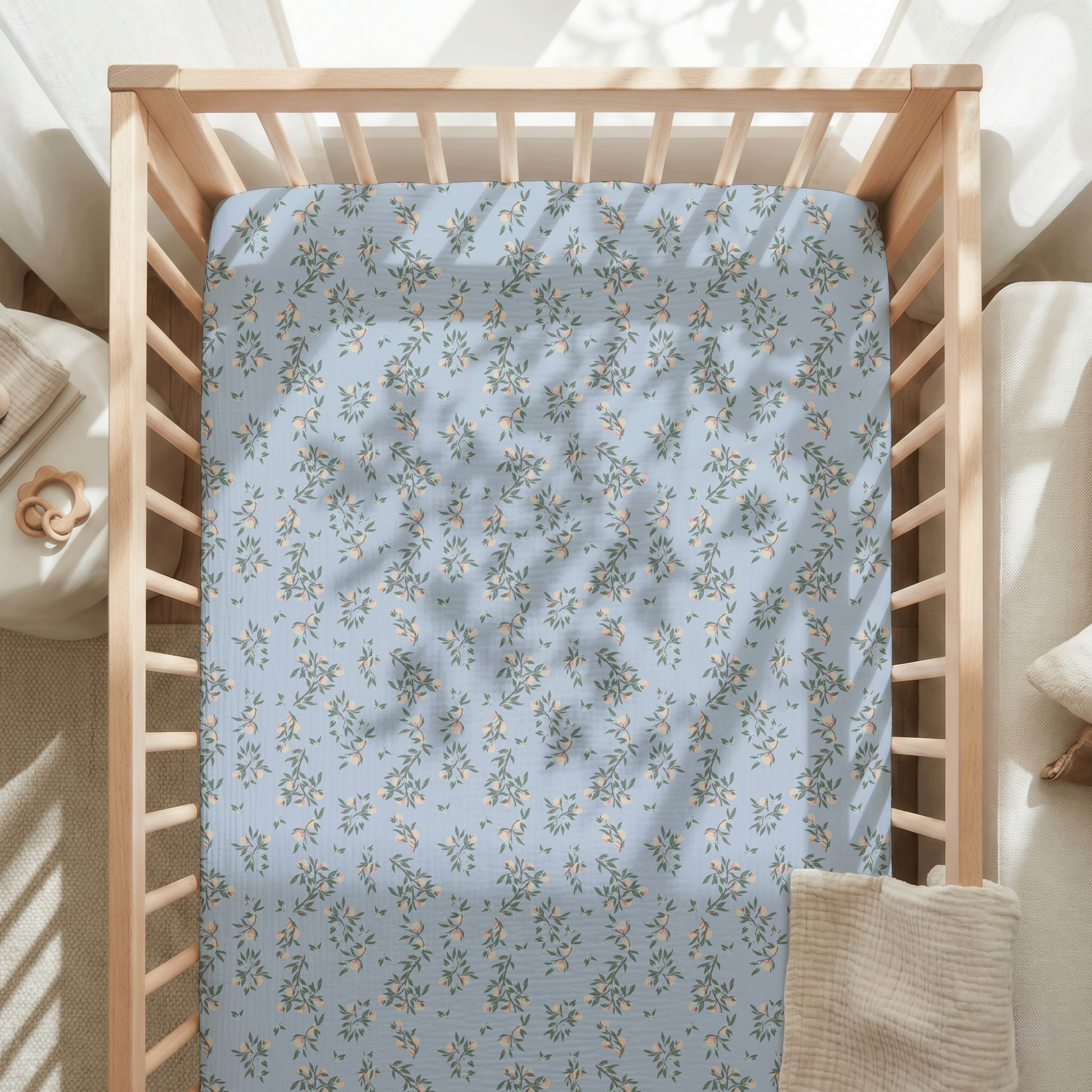Baby crib fitted sheet printed with small-scale floral surface pattern in soft blue and cream tones inside a light wood nursery setting.