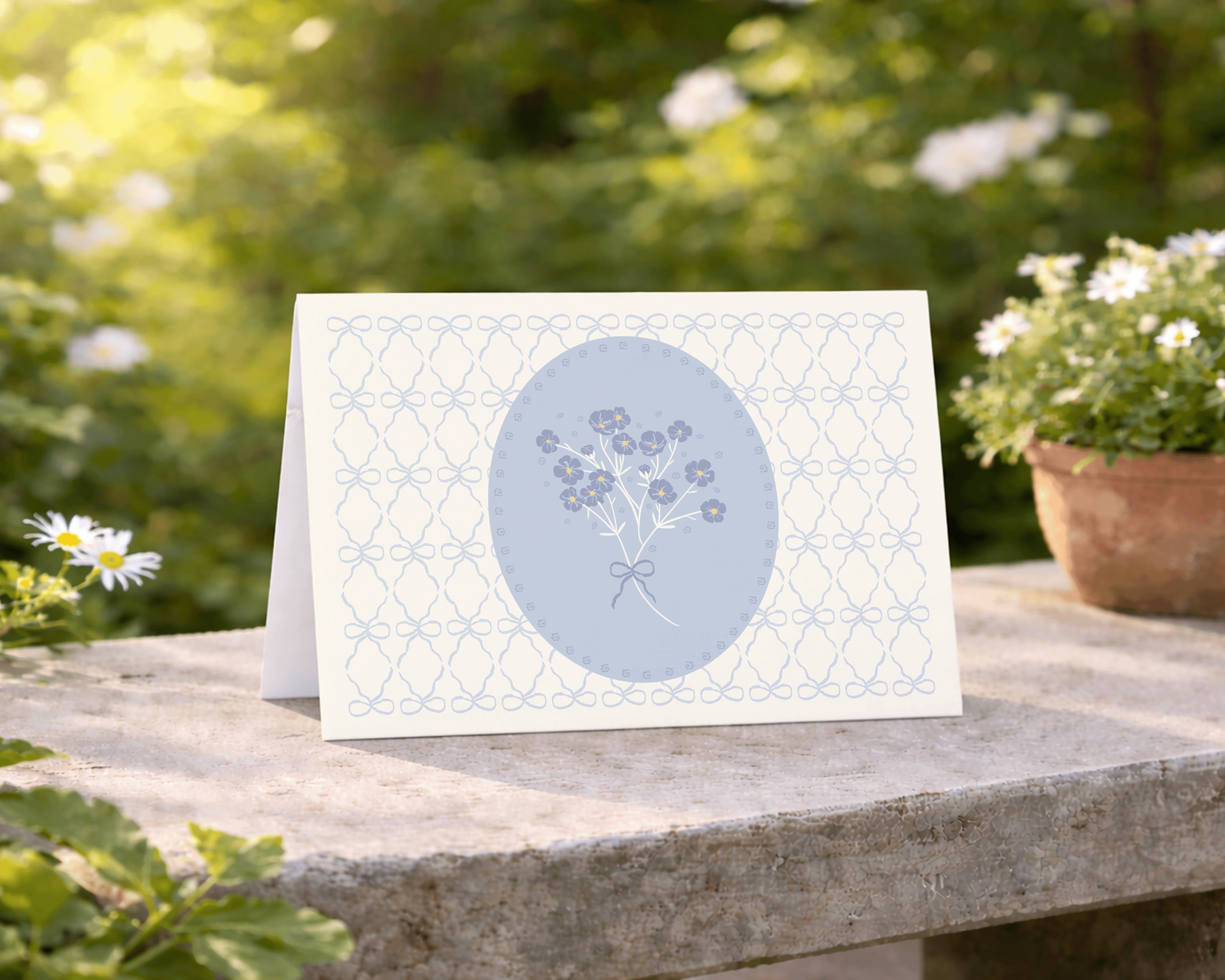 Greeting card featuring an engineered floral bouquet design in soft blue and cream, displayed on a stone table in a garden setting.