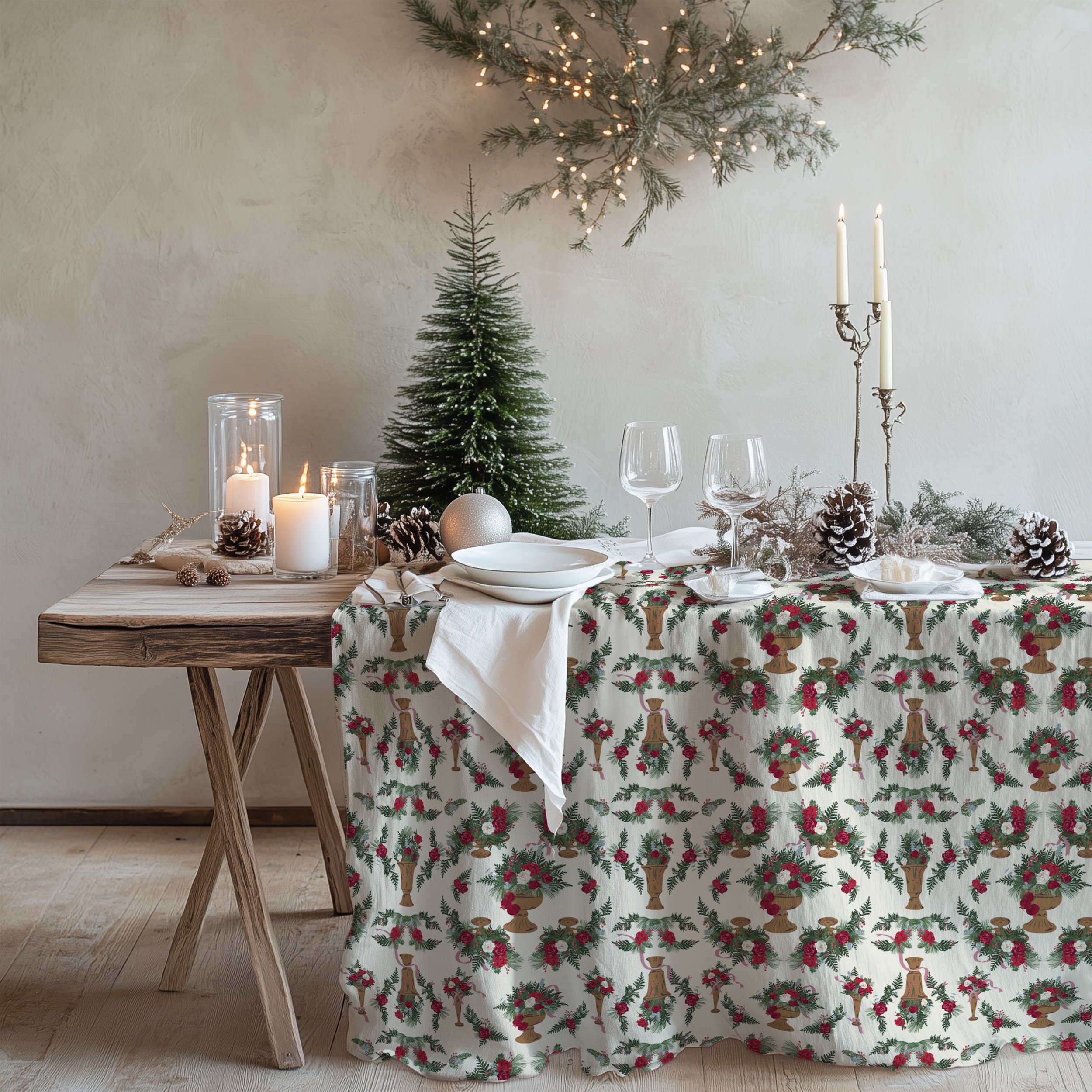 Rustic holiday table set with candles, evergreen branches, and dinnerware on a tablecloth printed with elegant winter floral arrangements in antique pedestal vessels.