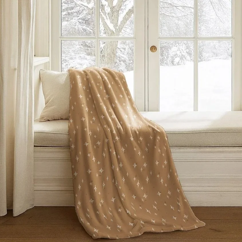 Cozy camel-colored throw blanket patterned with scattered cream stars, draped across a window seat overlooking a snowy winter landscape.