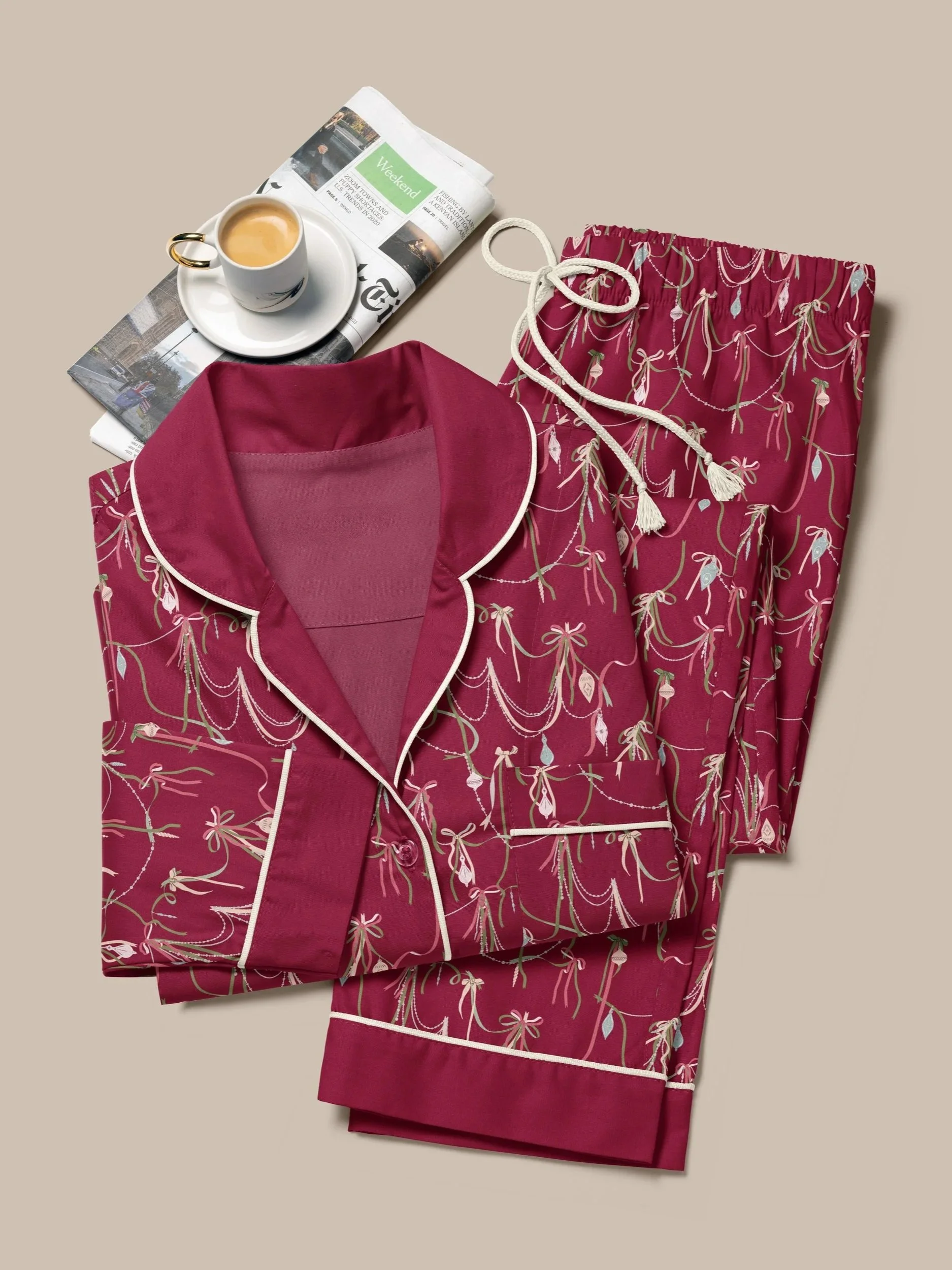 Folded cranberry-colored pajama set printed with draped velvet ribbons, glass ornaments, and delicate bead garlands, styled beside a cup of espresso and a folded newspaper.