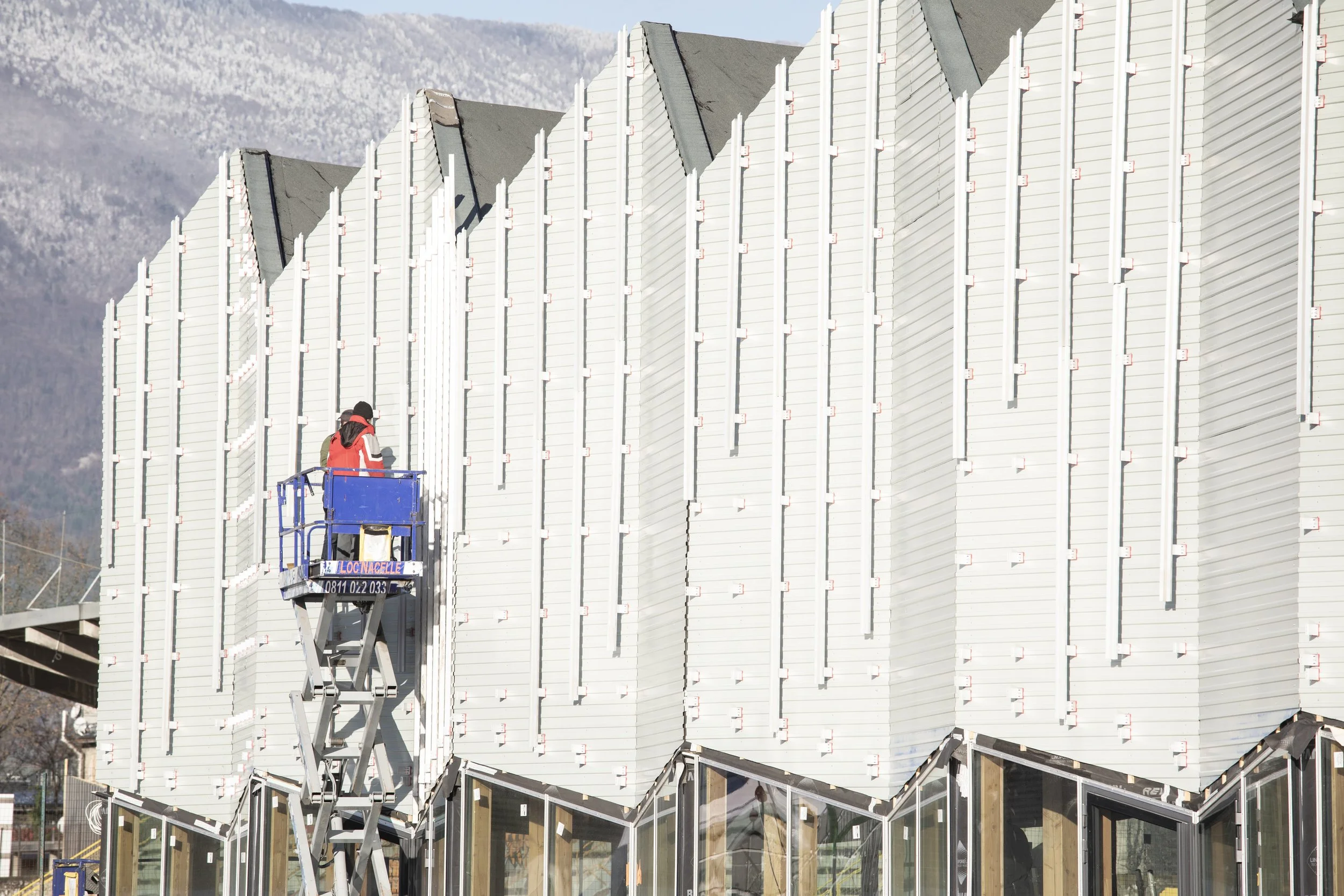 Travailleur sur une plateforme élévatrice en train d'installer ou de réparer la façade d'un bâtiment moderne avec des volets en lamelles verticales.