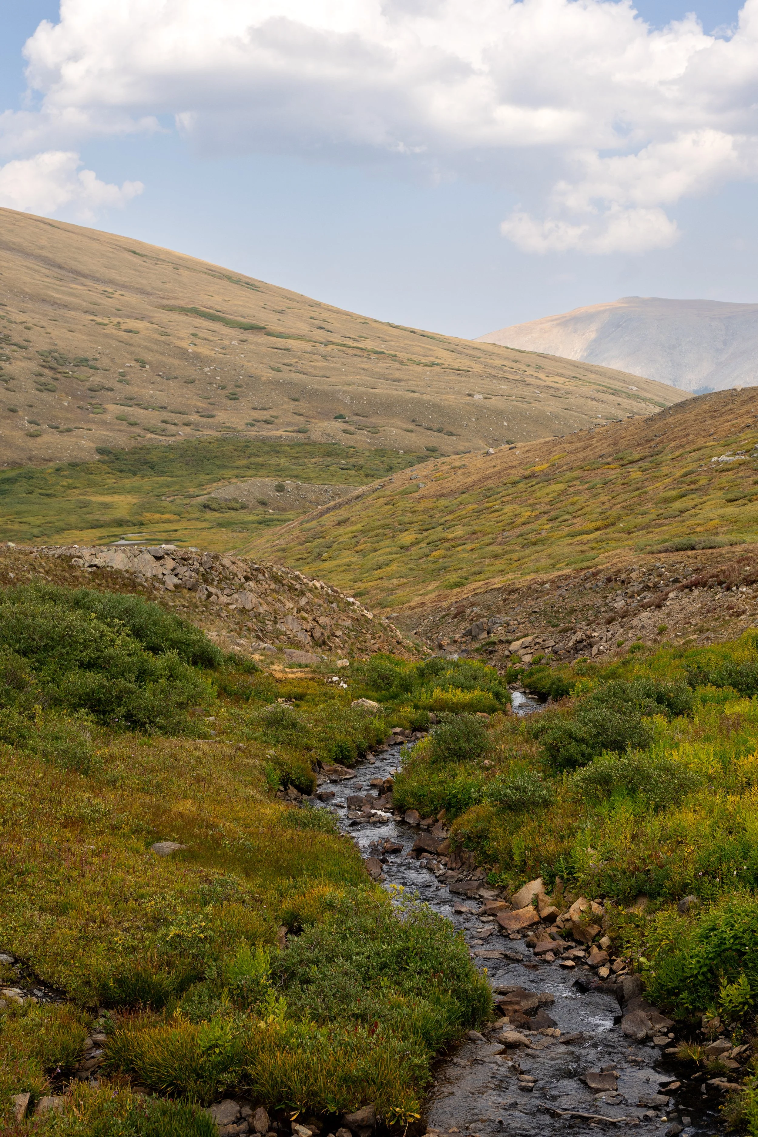 A mountain landscape with rolling hills, green vegetation, and a small stream flowing through the valley.