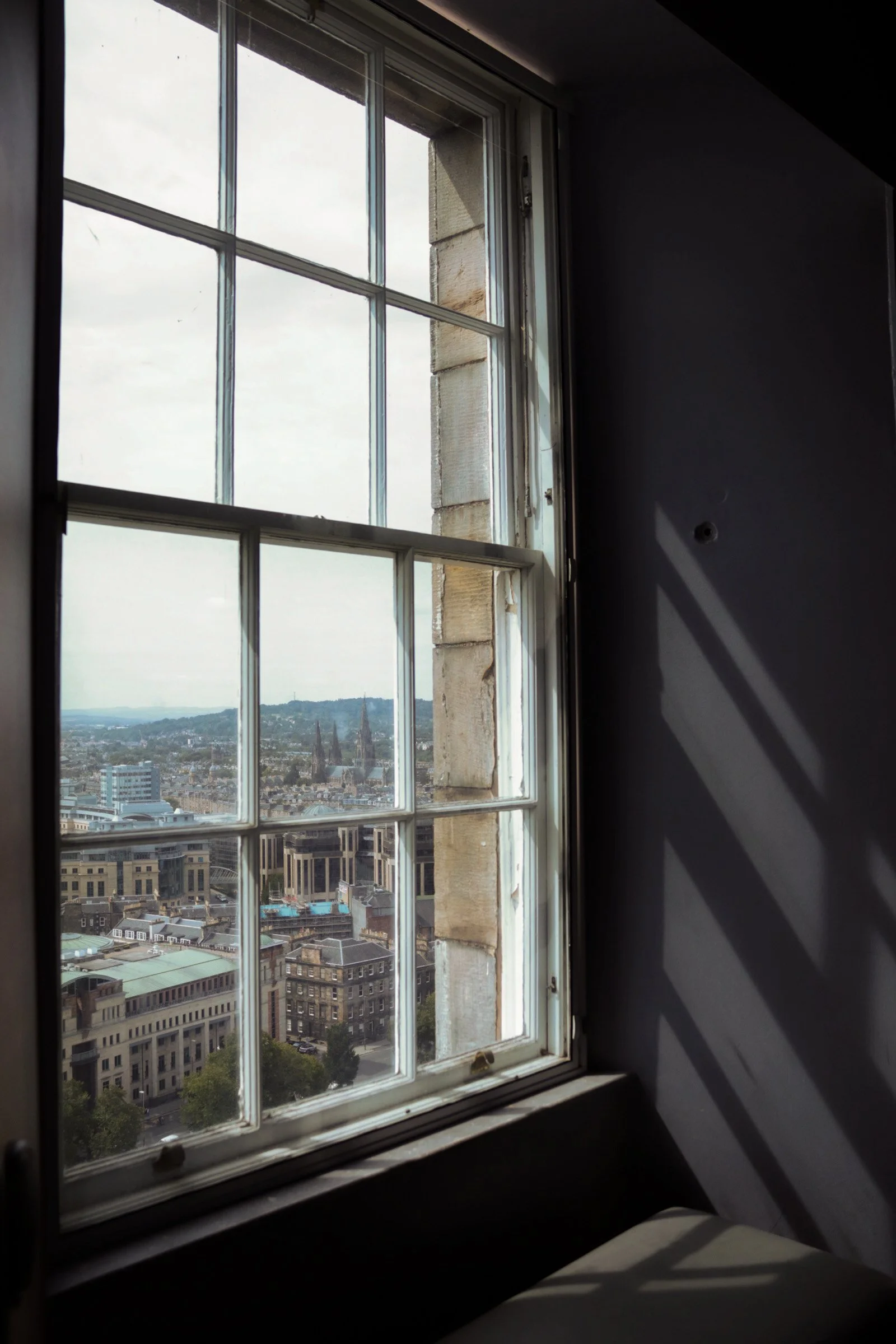 View through a window showing a cityscape with various buildings and a church spire, with shadows cast on an interior wall.
