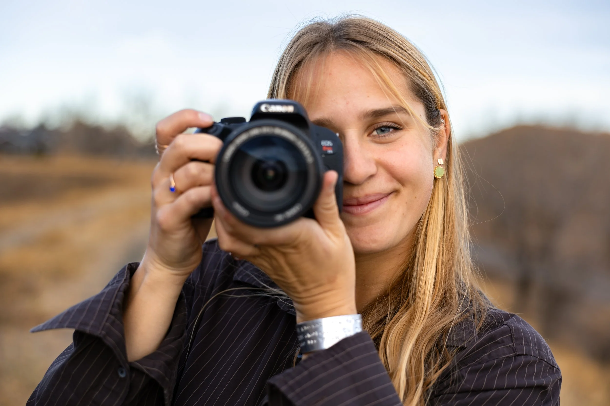 A woman with long blonde hair smiling while holding a Canon camera outdoors in a natural setting.