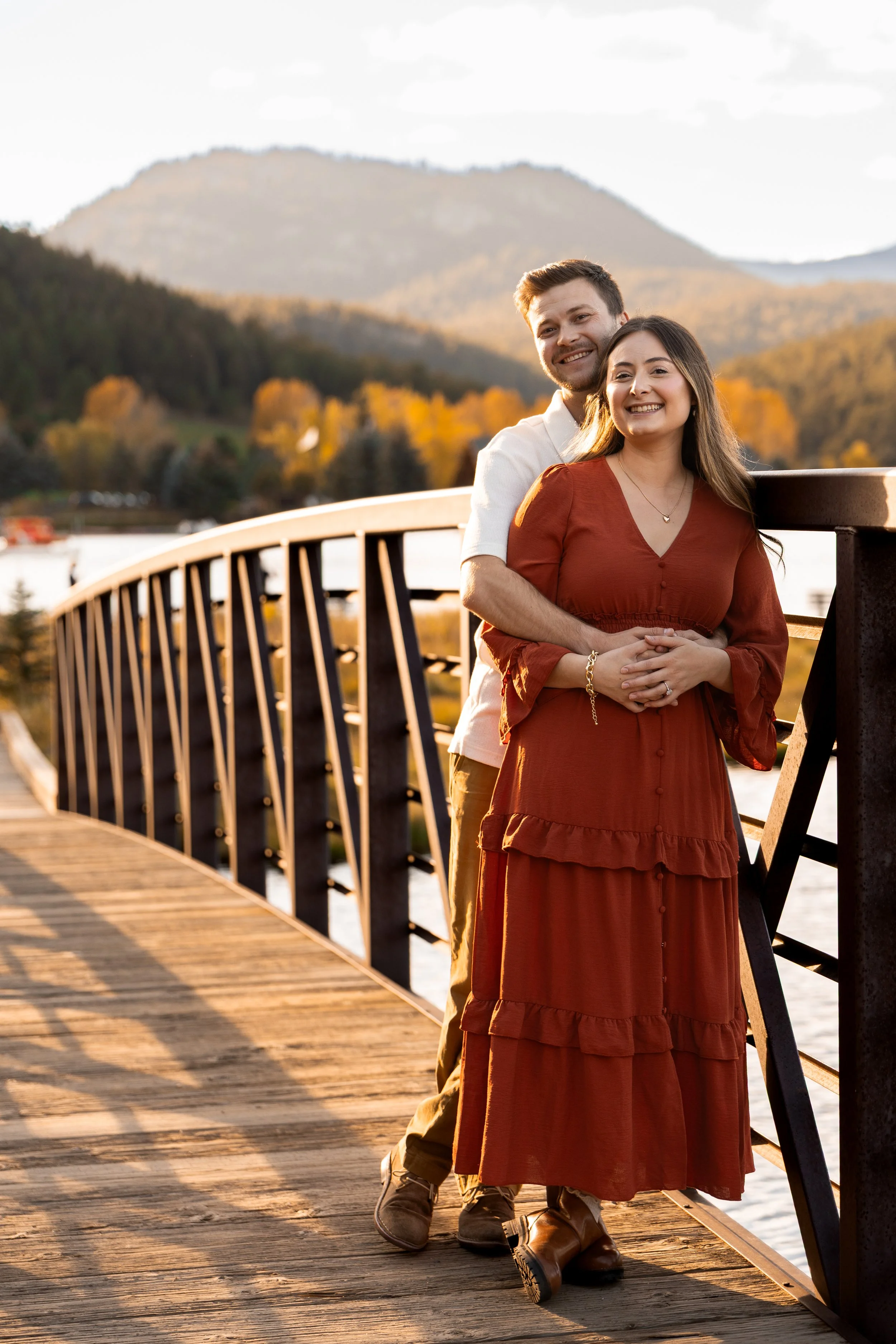 A smiling couple stands on a wooden bridge during sunset, with mountains and autumn trees in the background.