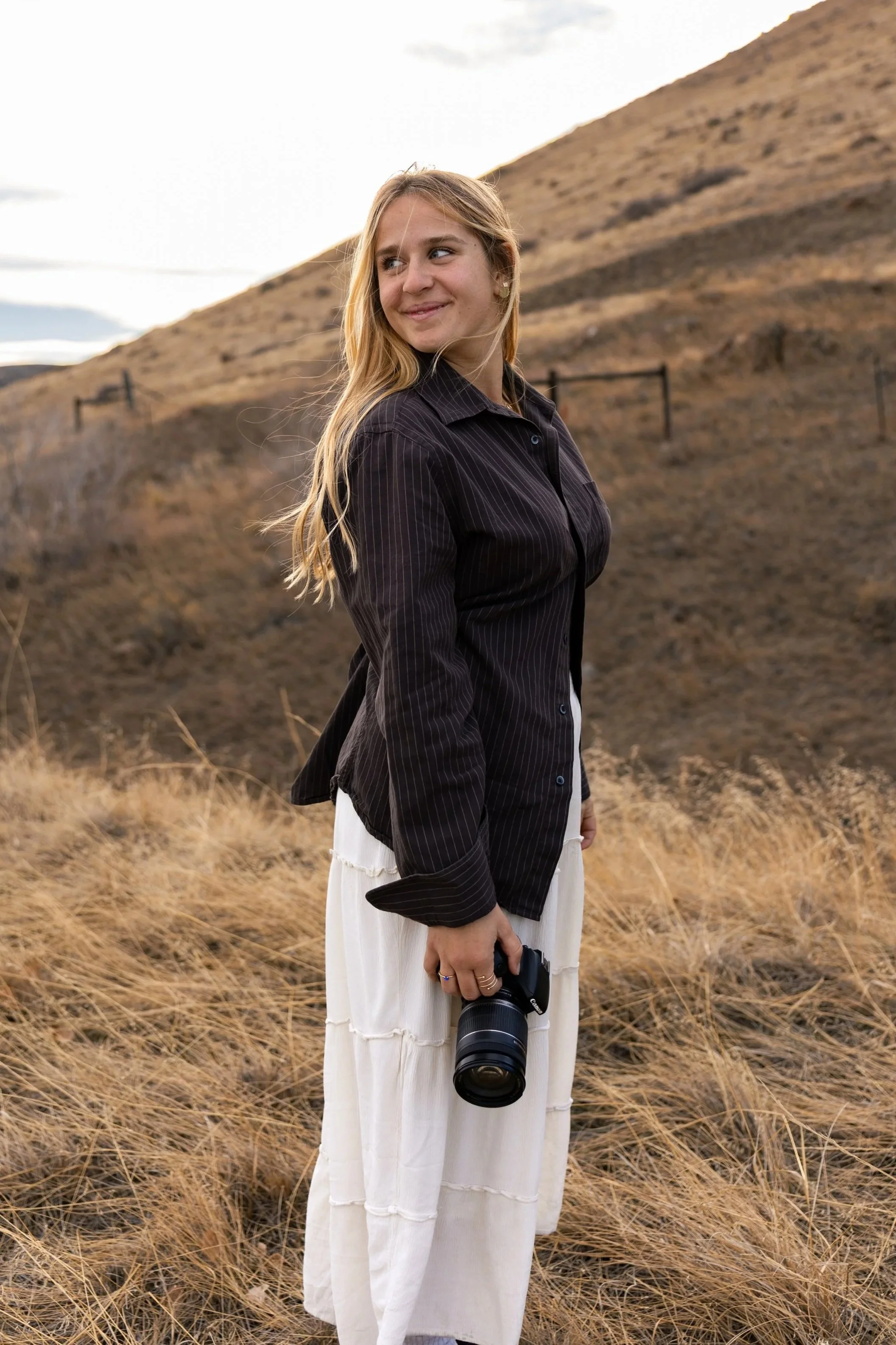 A young woman standing outdoors in a dry, grassy area holding a Canon camera in her right hand, with a hilly landscape in the background during the daytime.