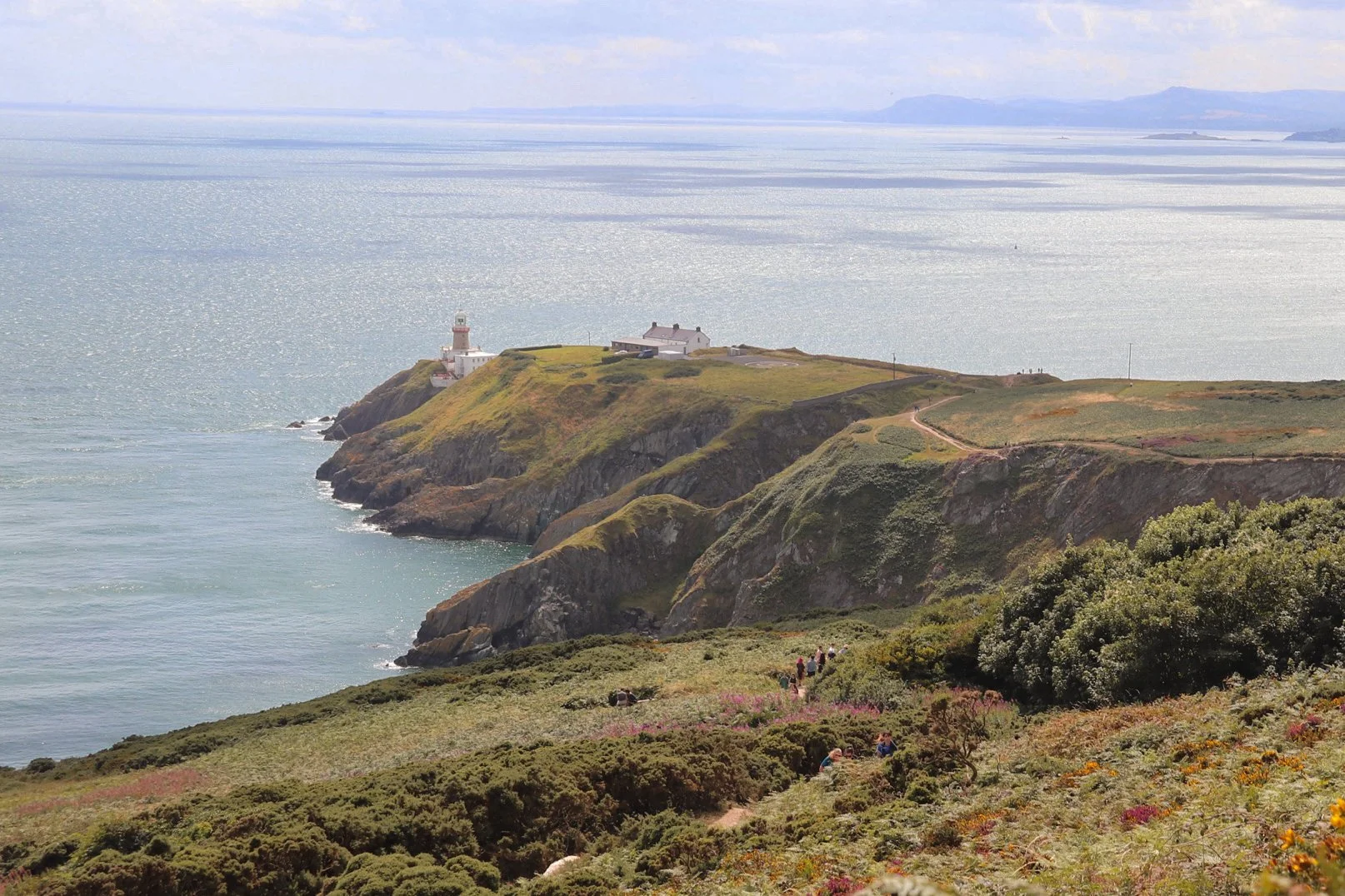 Cliffs along a coastline with a lighthouse and building at the top, overlooking the ocean, with a path and visitors in the foreground.