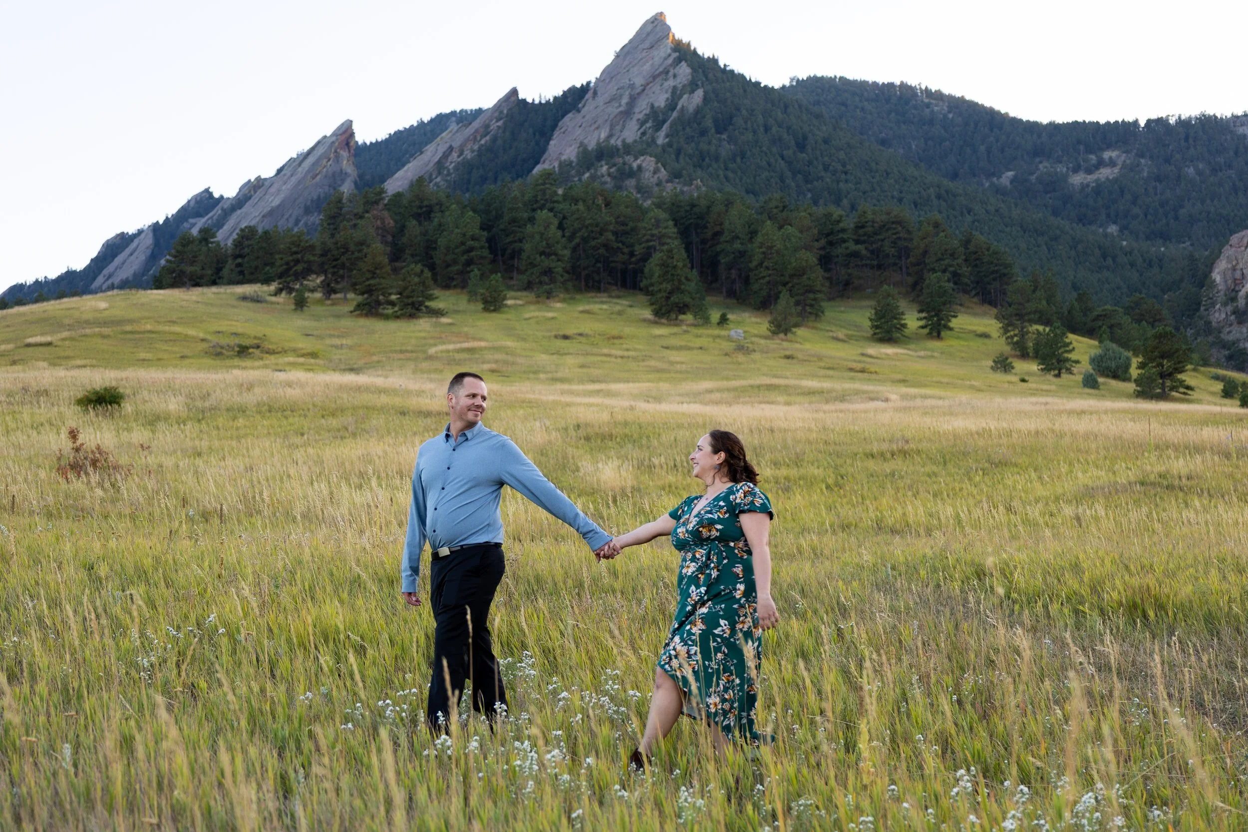 A couple holding hands in a grassy field with mountains and trees in the background.